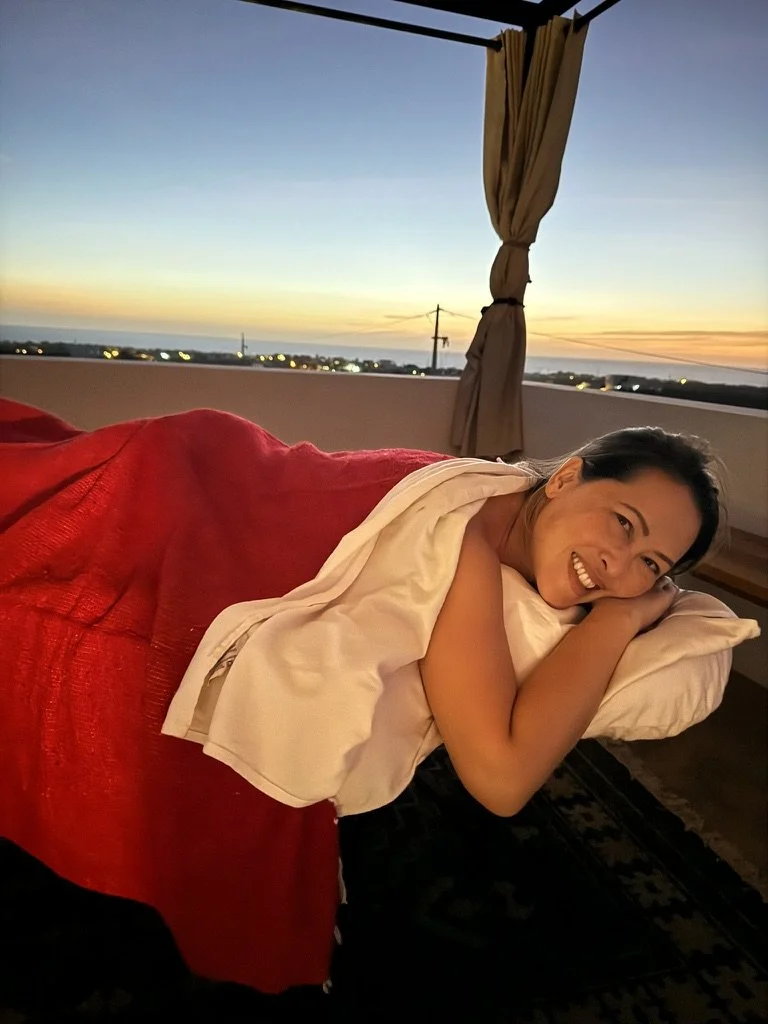 A woman lying on a bed, smiling, with a sunset view of the sky and distant city lights in the background.