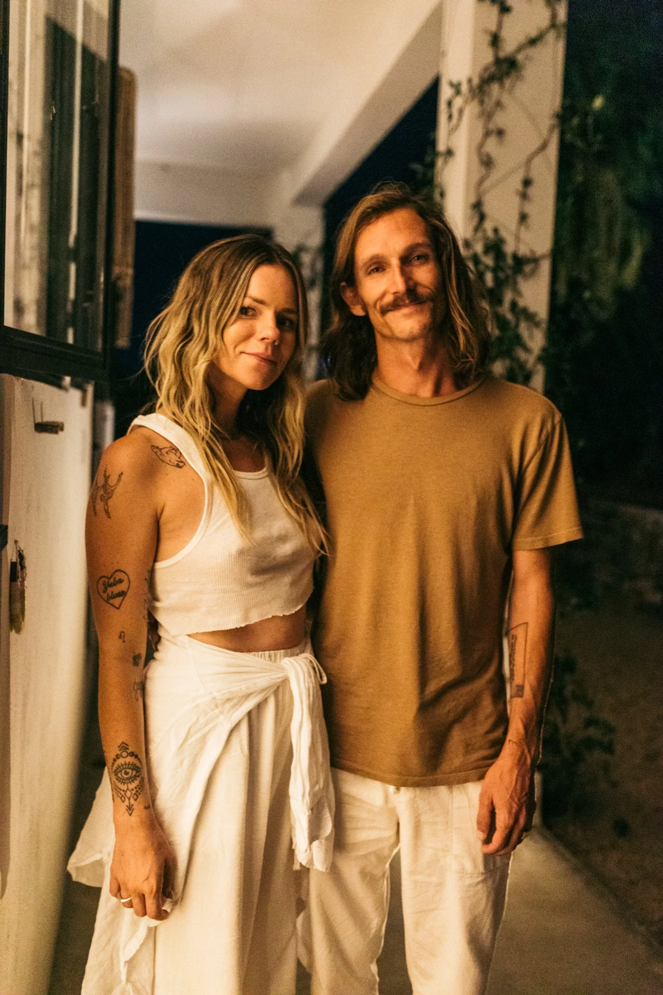 A young woman and a man pose together outside at night, standing near a wall and greenery, smiling at the camera.