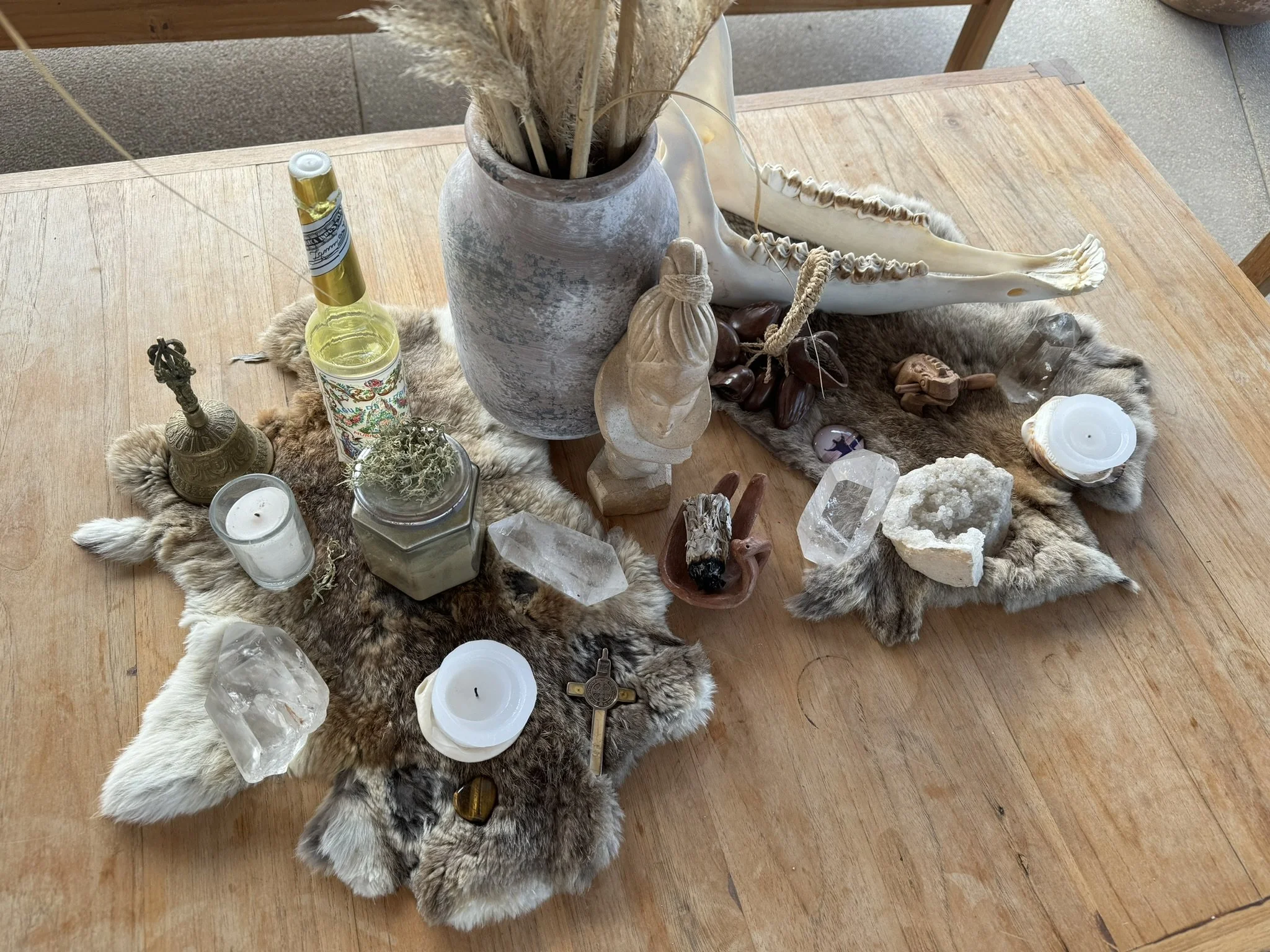 Arranged decorative objects on a fur and wood table, including crystals, candles, a small statue, a bottle, and dried plants.