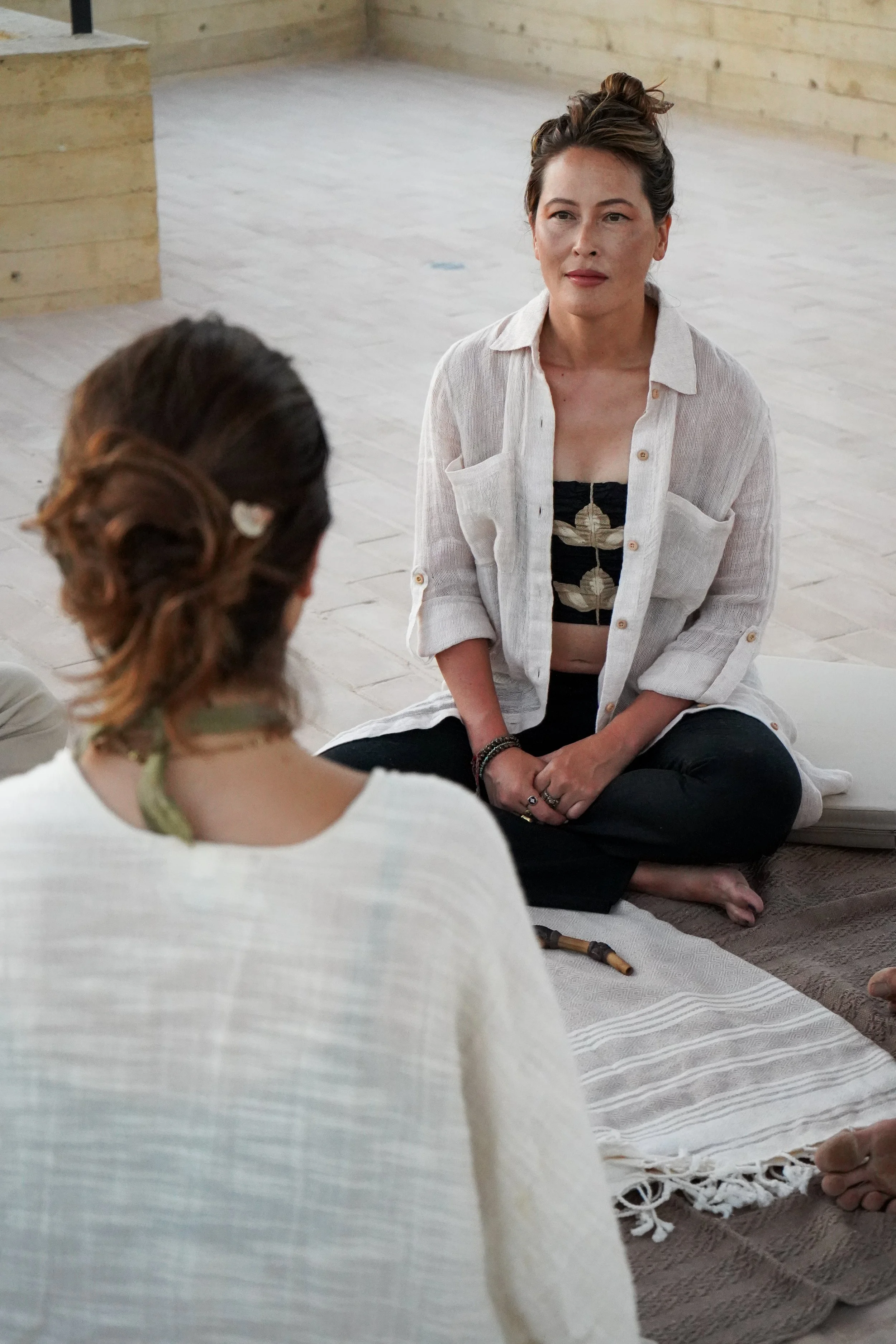 Two women sitting cross-legged on a rug, engaged in a conversation in a room with a wooden floor and wall. The woman in focus has dark brown hair styled in a bun and is wearing a sheer white shirt over a black top with leaf print, black pants, and several rings and bracelets.