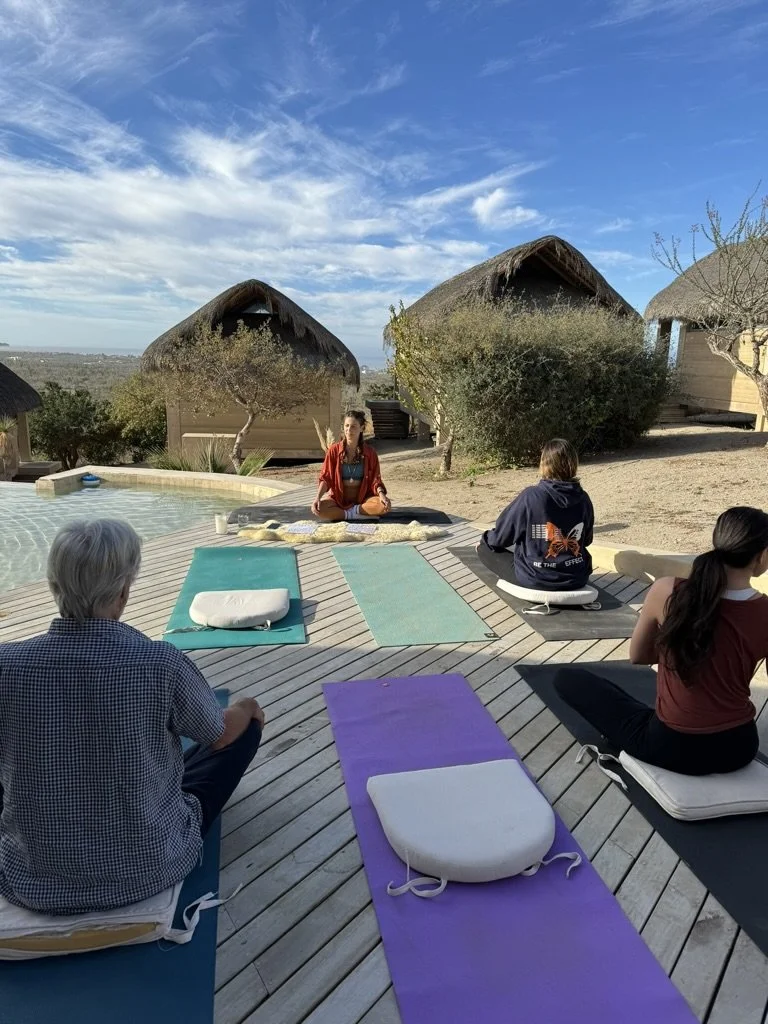 A group of people practicing yoga outdoors on wooden deck with thatched roof structures and blue sky in the background.