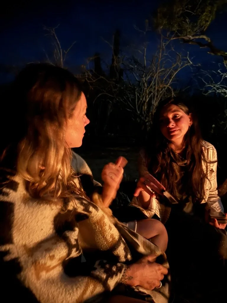 Two women sitting outdoors at night near a campfire, talking and holding cards, with a dark sky and trees in the background.