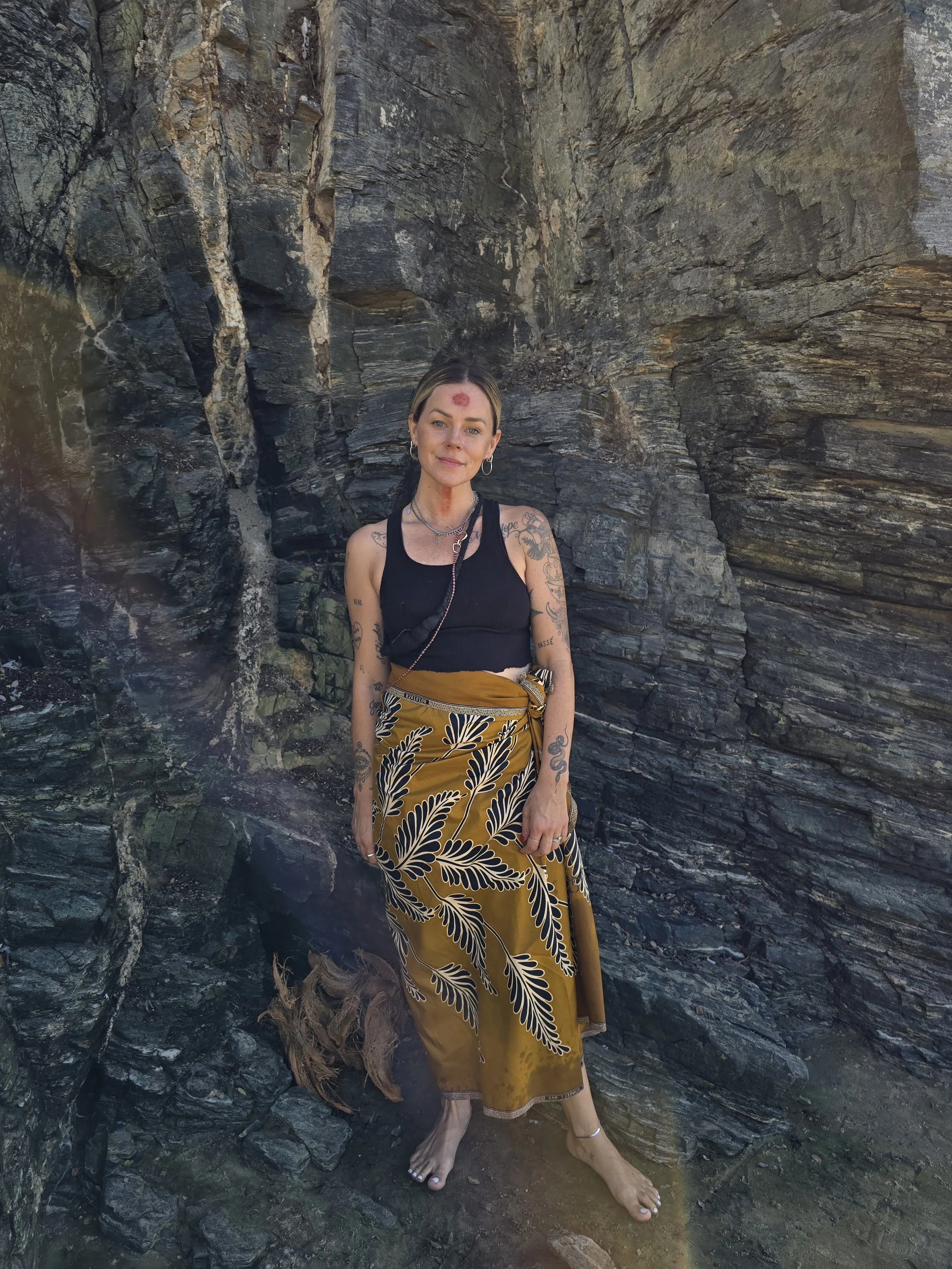 Woman standing barefoot on rocky terrain in front of a rugged rock wall, wearing a black tank top and a yellow skirt with black leaf patterns, with tattoos on her arms and face paint on her forehead.