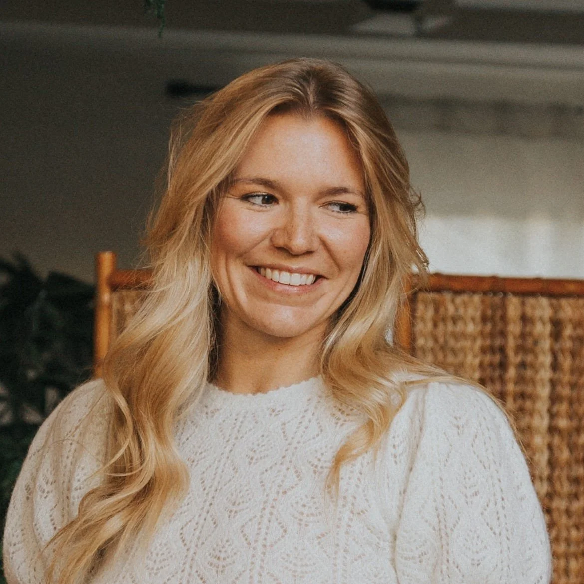 A woman with long blonde hair smiling, wearing a white knitted sweater, sitting indoors with a wicker chair and plants in the background.