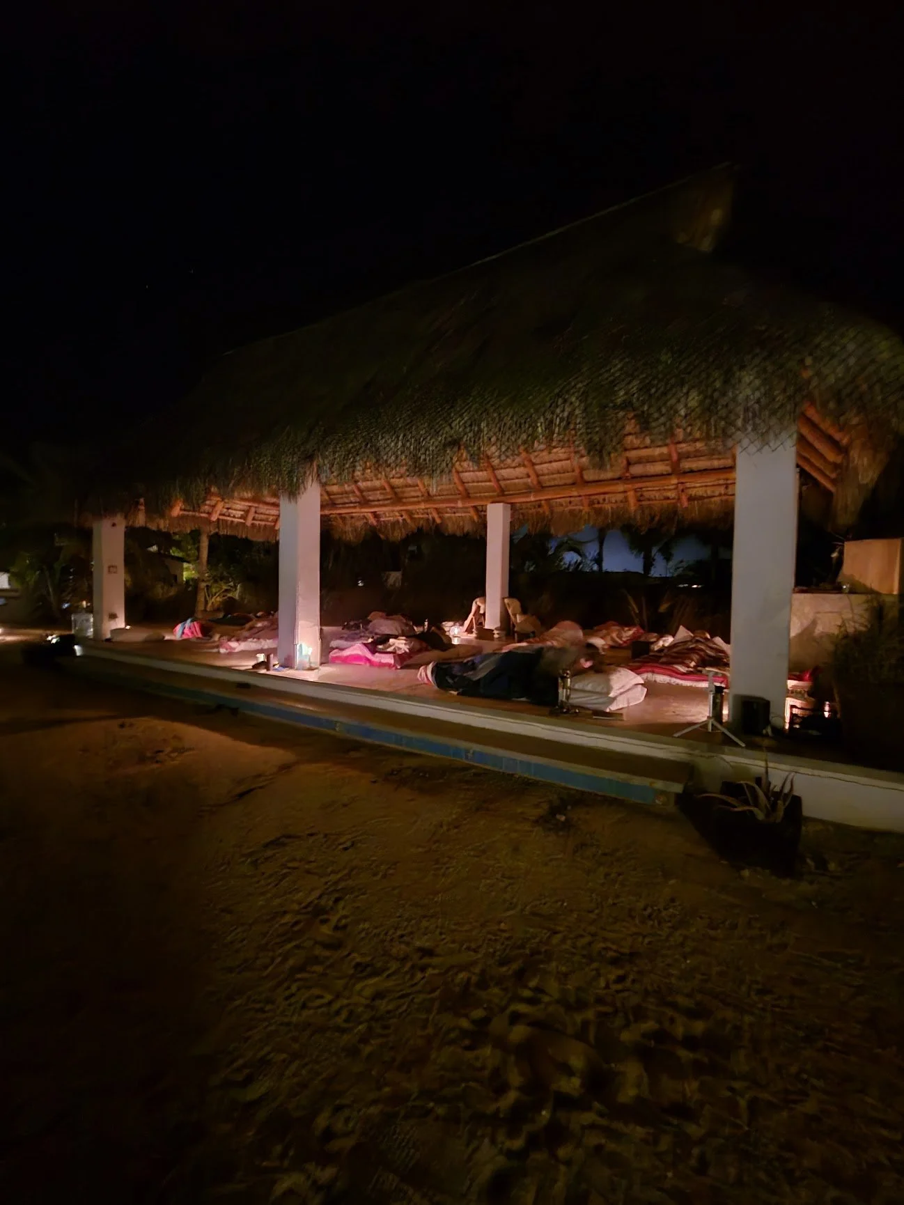 Nighttime scene of a thatched-roof open-air massage or relaxation area with multiple beds, some with people lying down, illuminated by soft lighting.