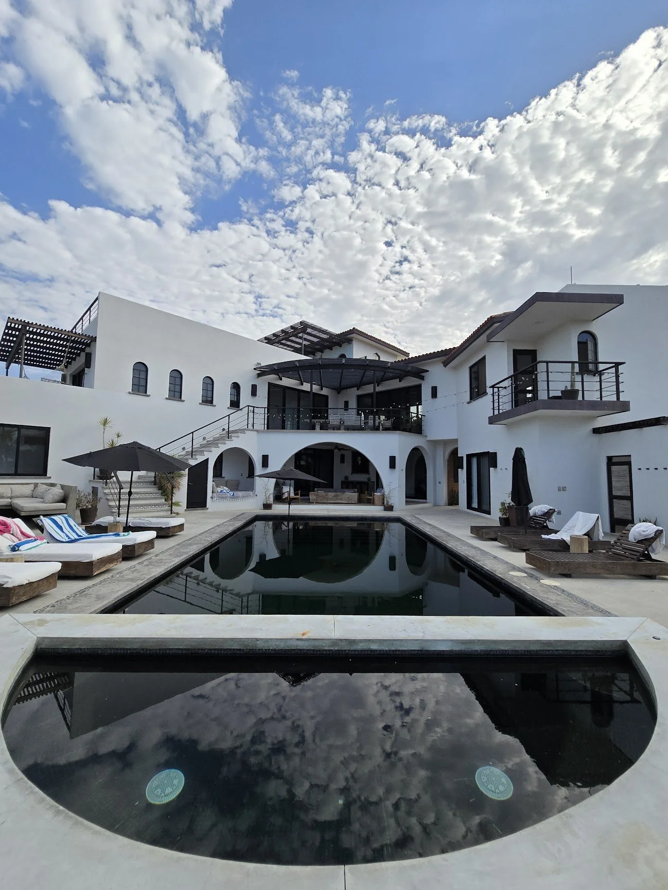 A modern white house with a pool and a hot tub, outdoor lounge chairs, umbrellas, and a cloudy sky.