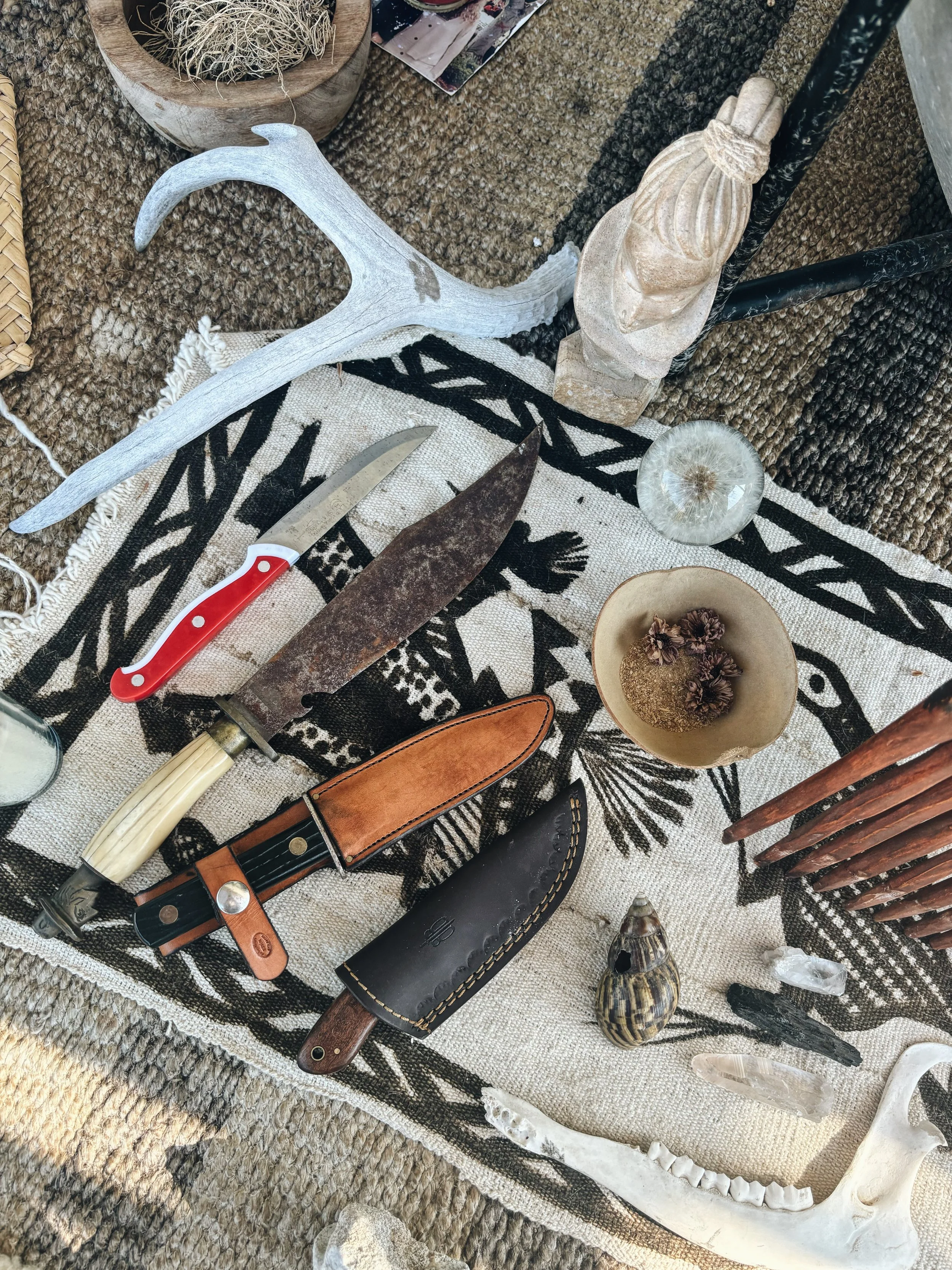 Collection of tribal artifacts and tools on a patterned cloth, including a large antler, a rusty knife, a smaller knife with a red handle, a leather sheath, a carved wooden sculpture, shells, a bone or horn, and a skull, surrounded by woven mats and natural elements.