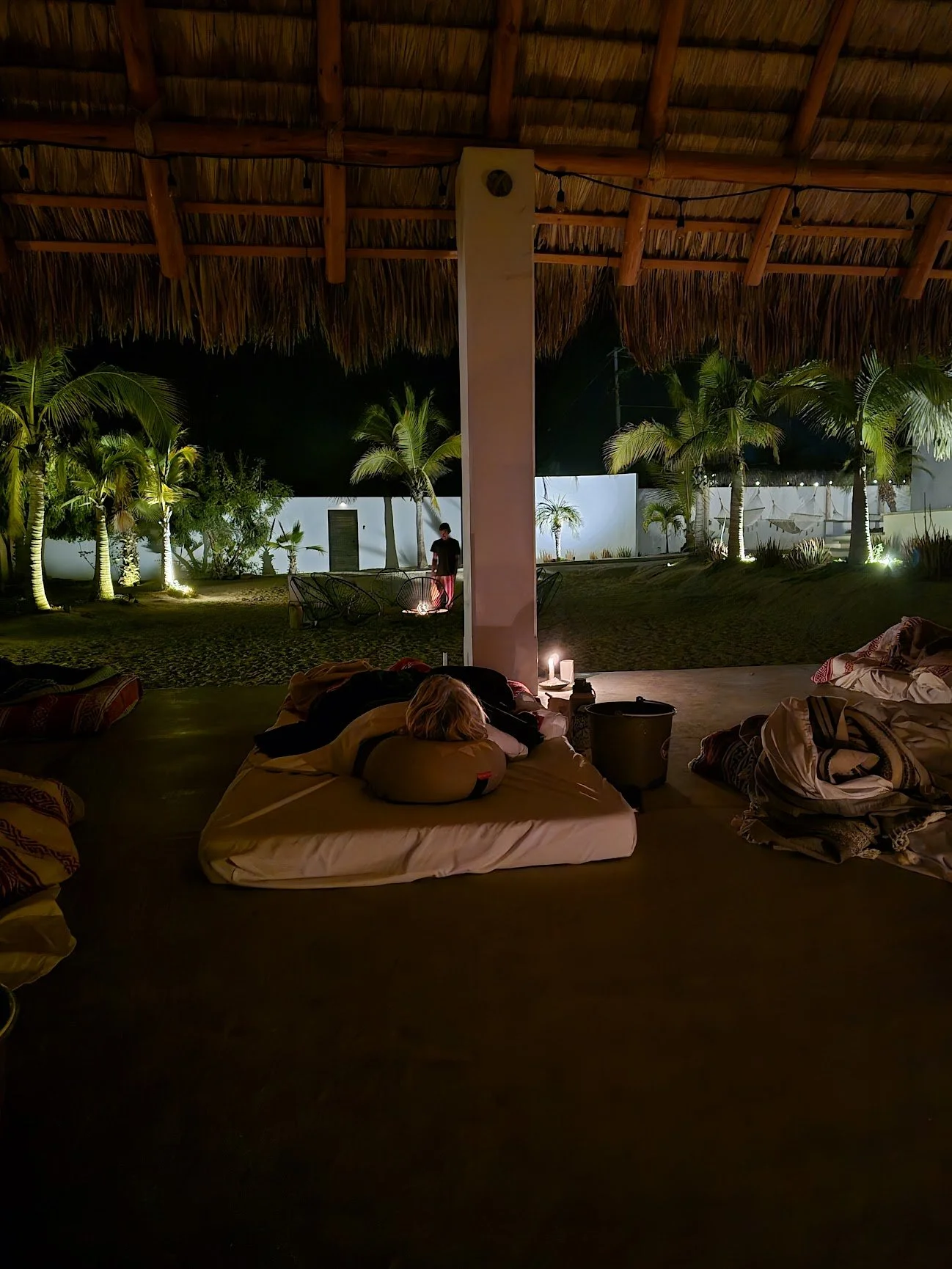 Nighttime view from a covered outdoor area with a thatched roof, showing a person sleeping on a mattress with a neck pillow, surrounded by scattered clothes and a bucket, looking out onto a lit garden with palm trees and a person standing outside.