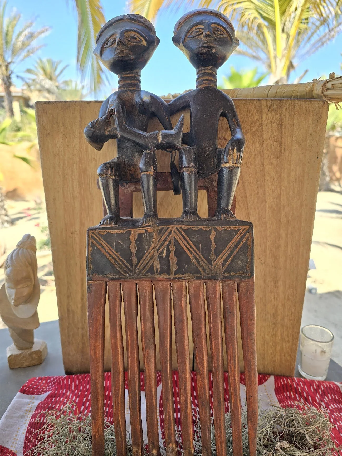 Wooden tribal sculpture of two figures sitting back-to-back on a carved base, with palm trees and a blue sky in the background.