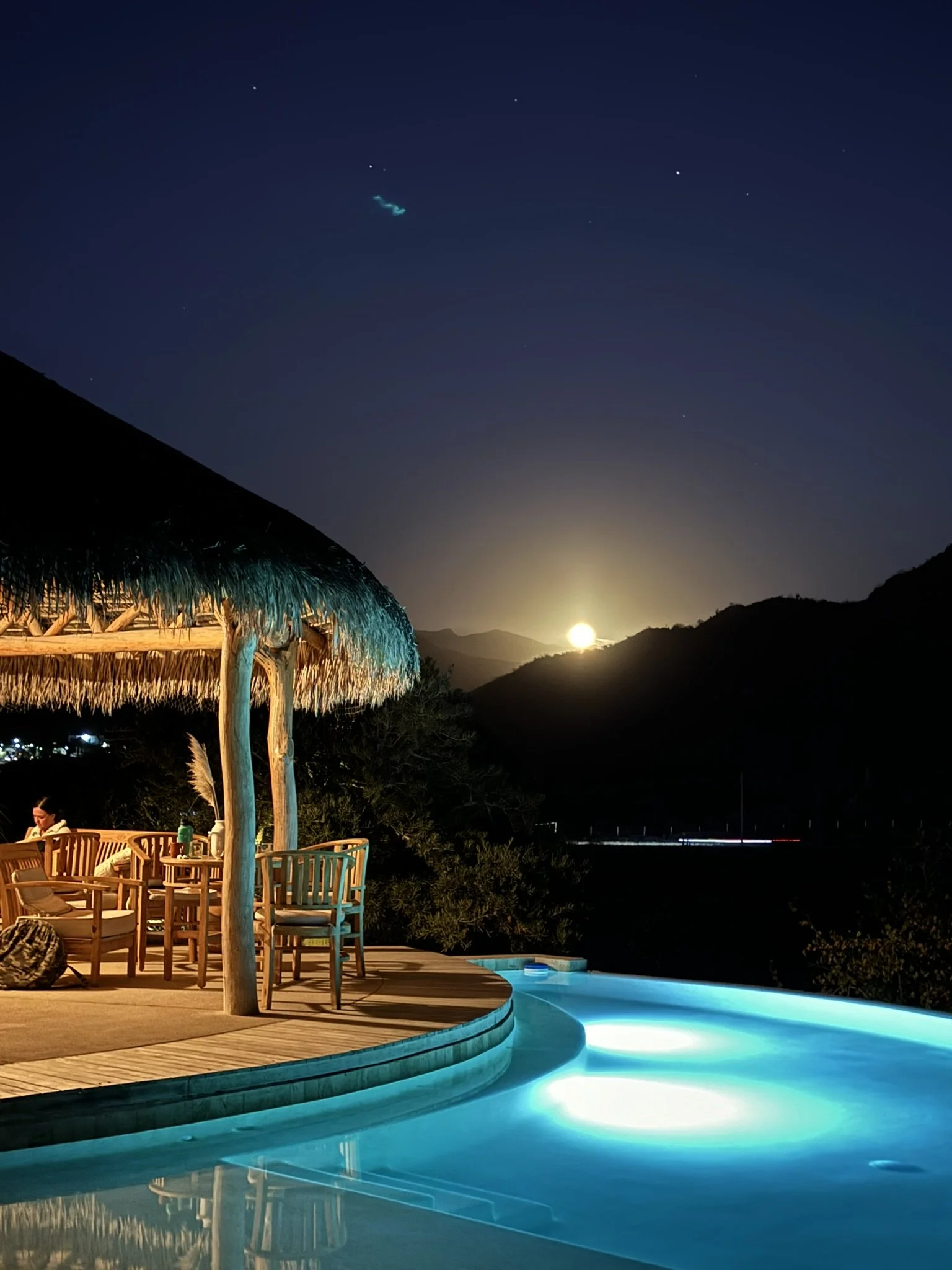Nighttime scene with a lit swimming pool, a thatched-roof cabana, mountain silhouette, and a bright full moon in the sky.