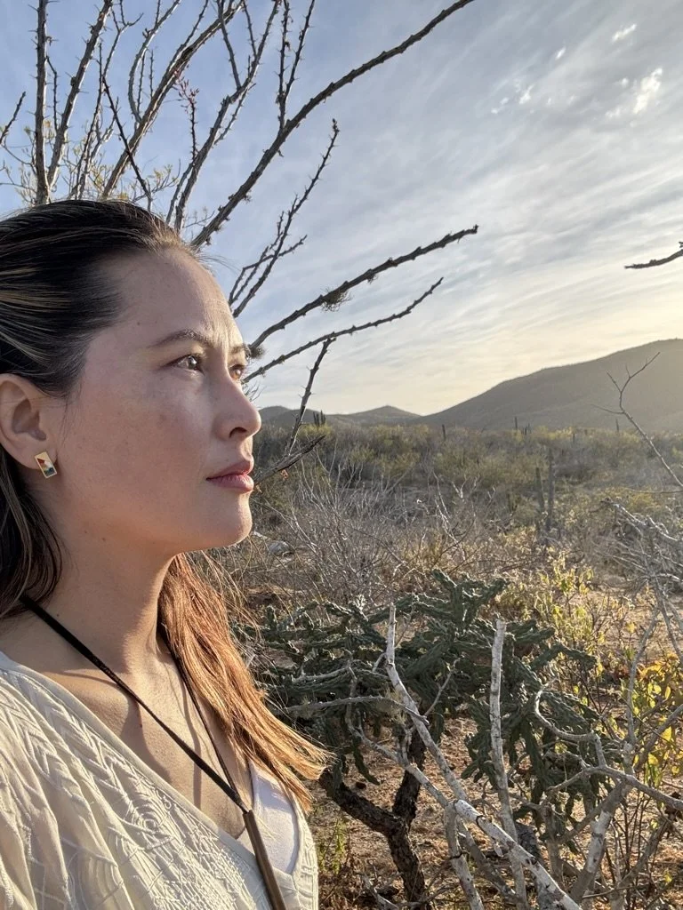 Close-up of a woman with long brown hair, wearing earrings and a white top, standing in a desert landscape with sparse vegetation and mountains in the background, during sunset.