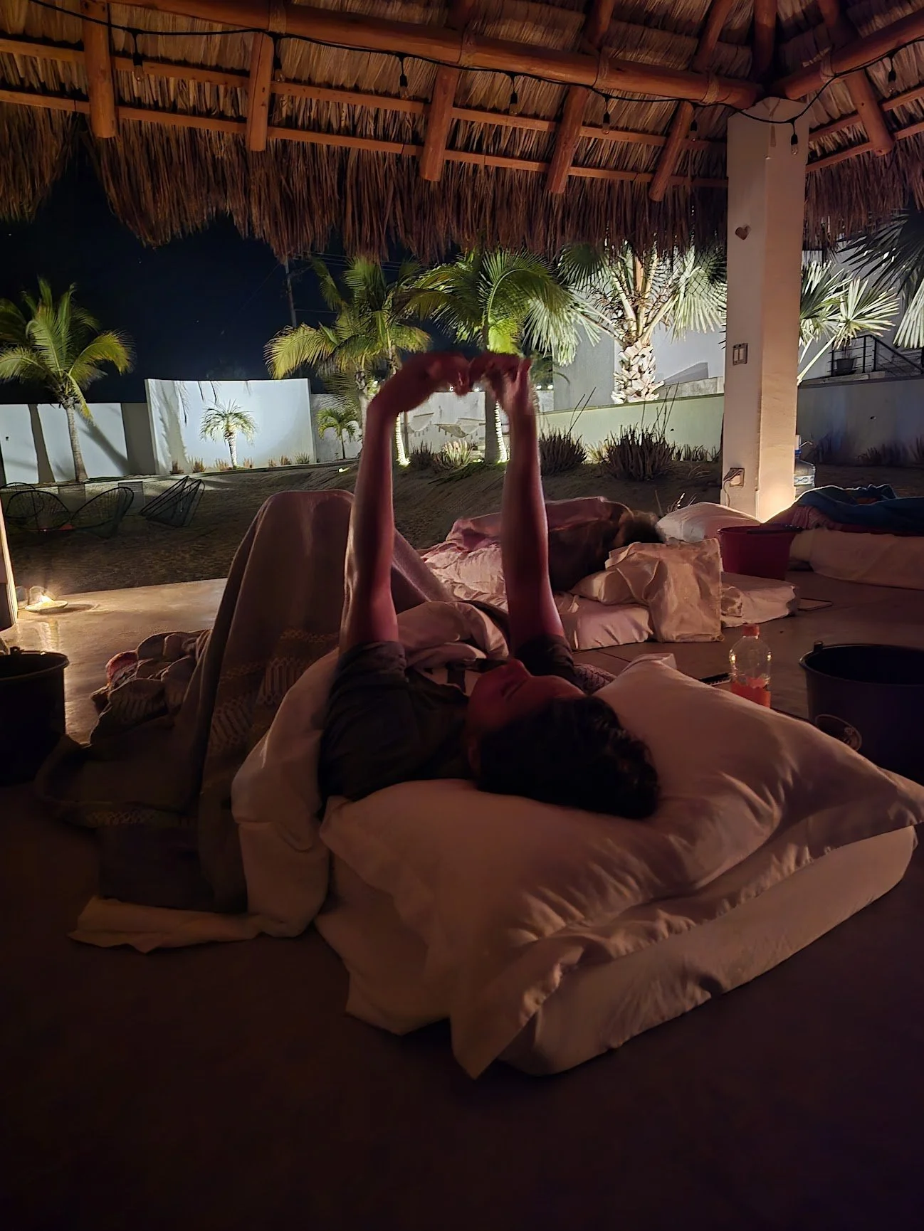Person lying on their back in a bed under a thatched roof, stretching their arms overhead, with palm trees and outdoor scenery visible through open sides at night.