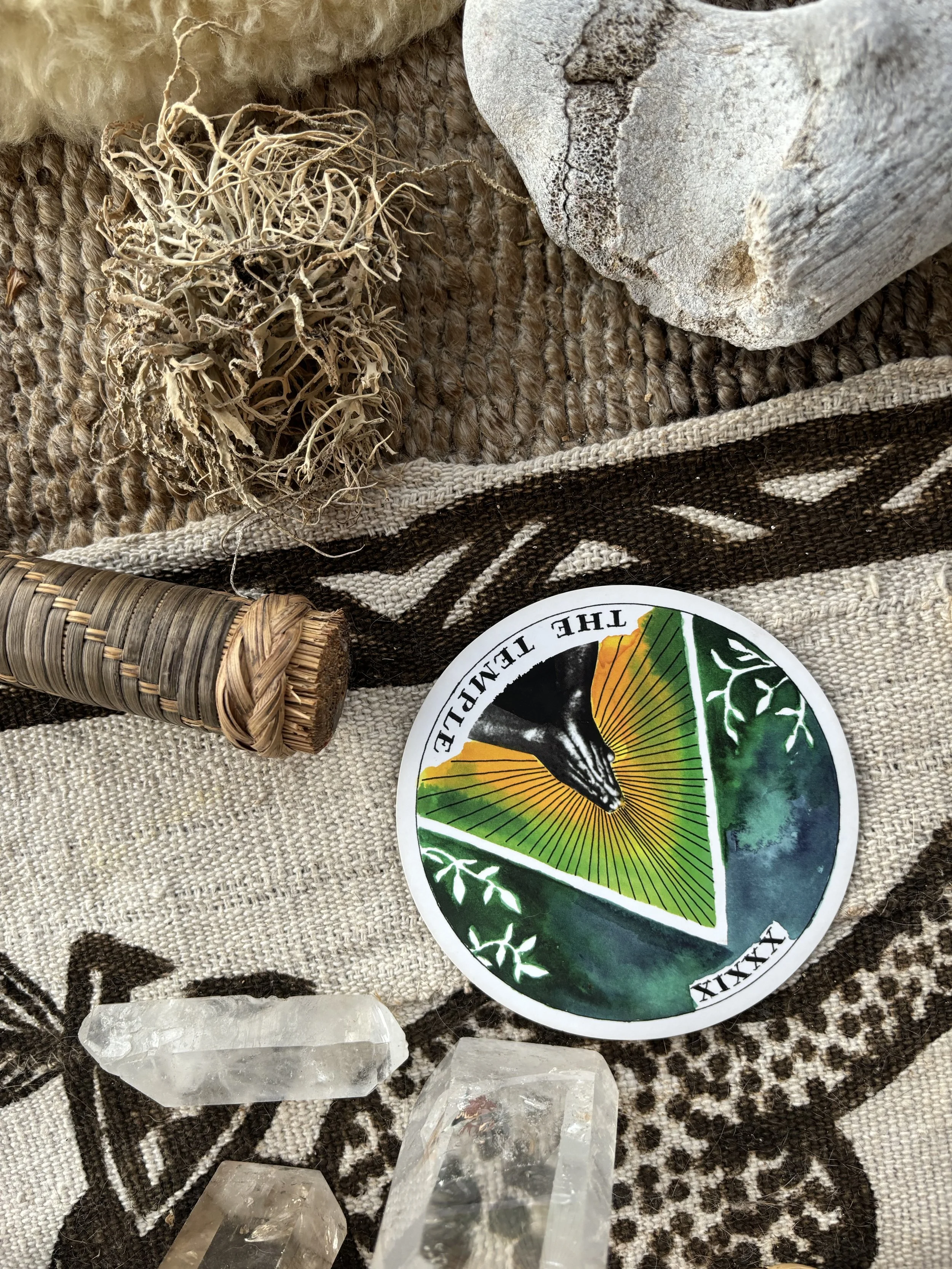 A collection of spiritual and natural objects on a textured cloth, including a terrarium plant, a large stone, a stick wrapped with woven material, a circular sticker with a black panther and the words "THE TEMPLE" and Roman numeral "XXIX," and multiple clear quartz crystals.