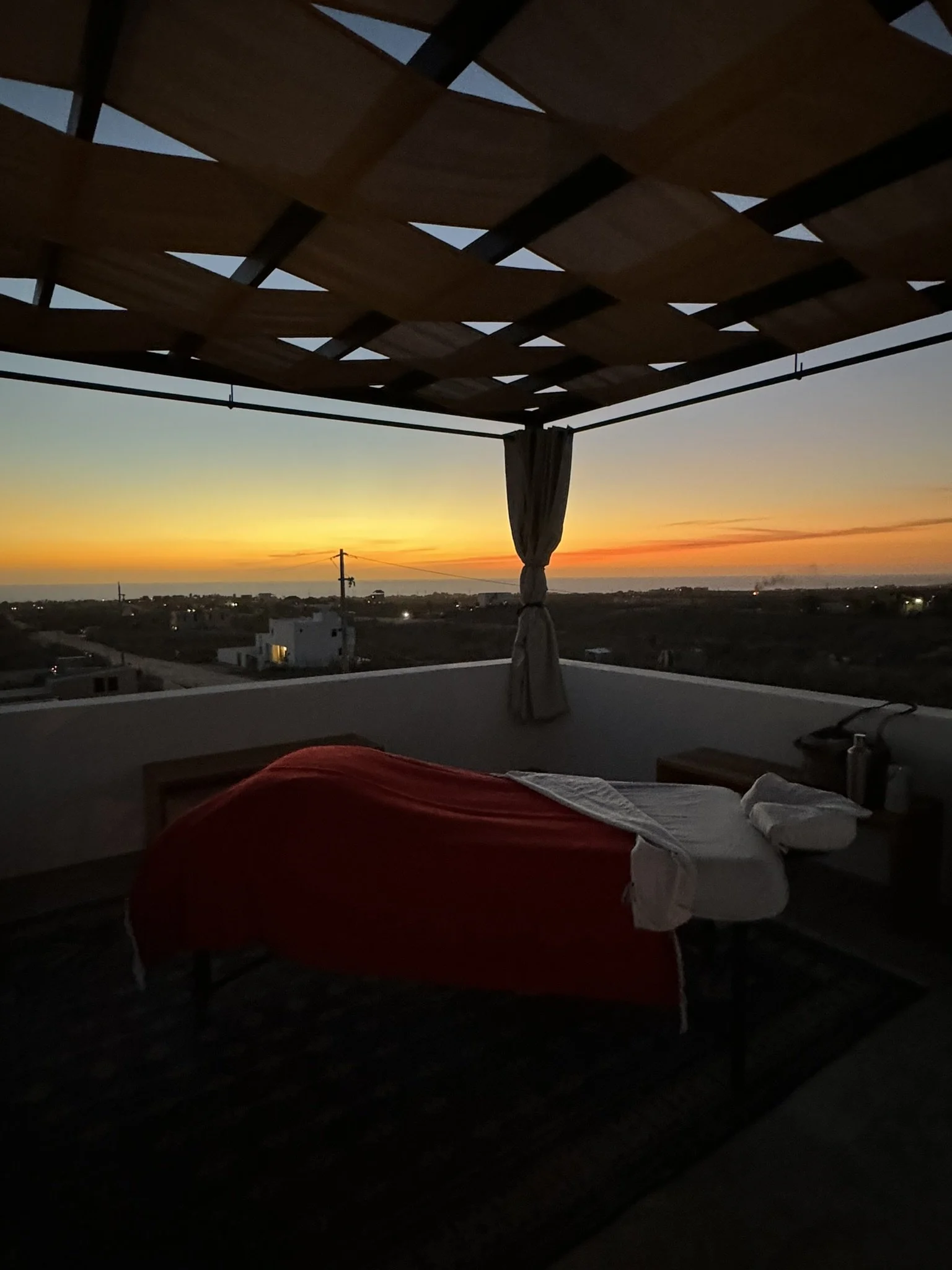 Outdoor rooftop scene during sunset with a bed covered in a red blanket and a white sheet, a small table with a water bottle, and an open roof structure made of wood and fabric panels.