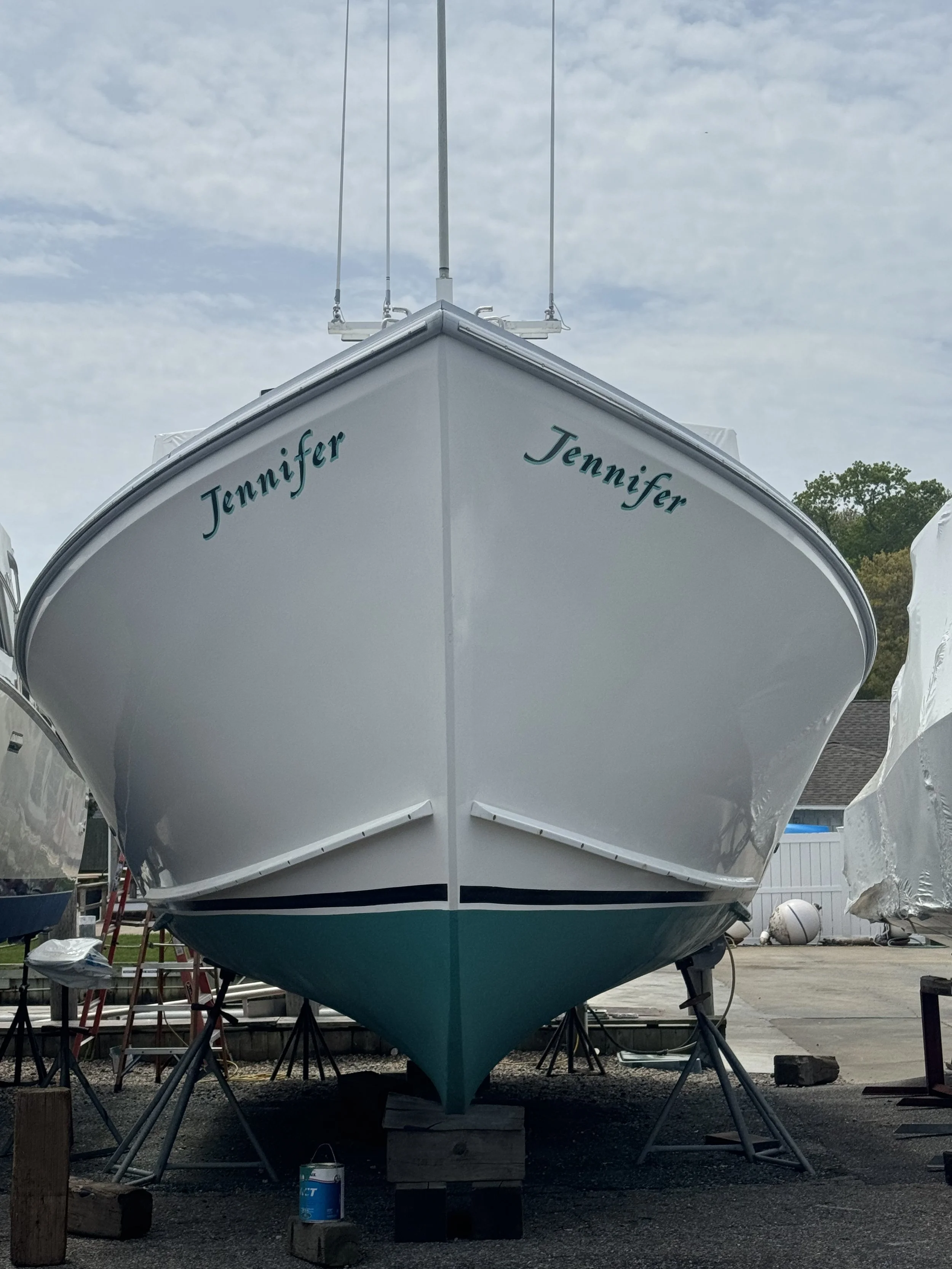 A white boat named Jennifer is on display on land, supported by stands and blocks, with a partly cloudy sky in the background.
