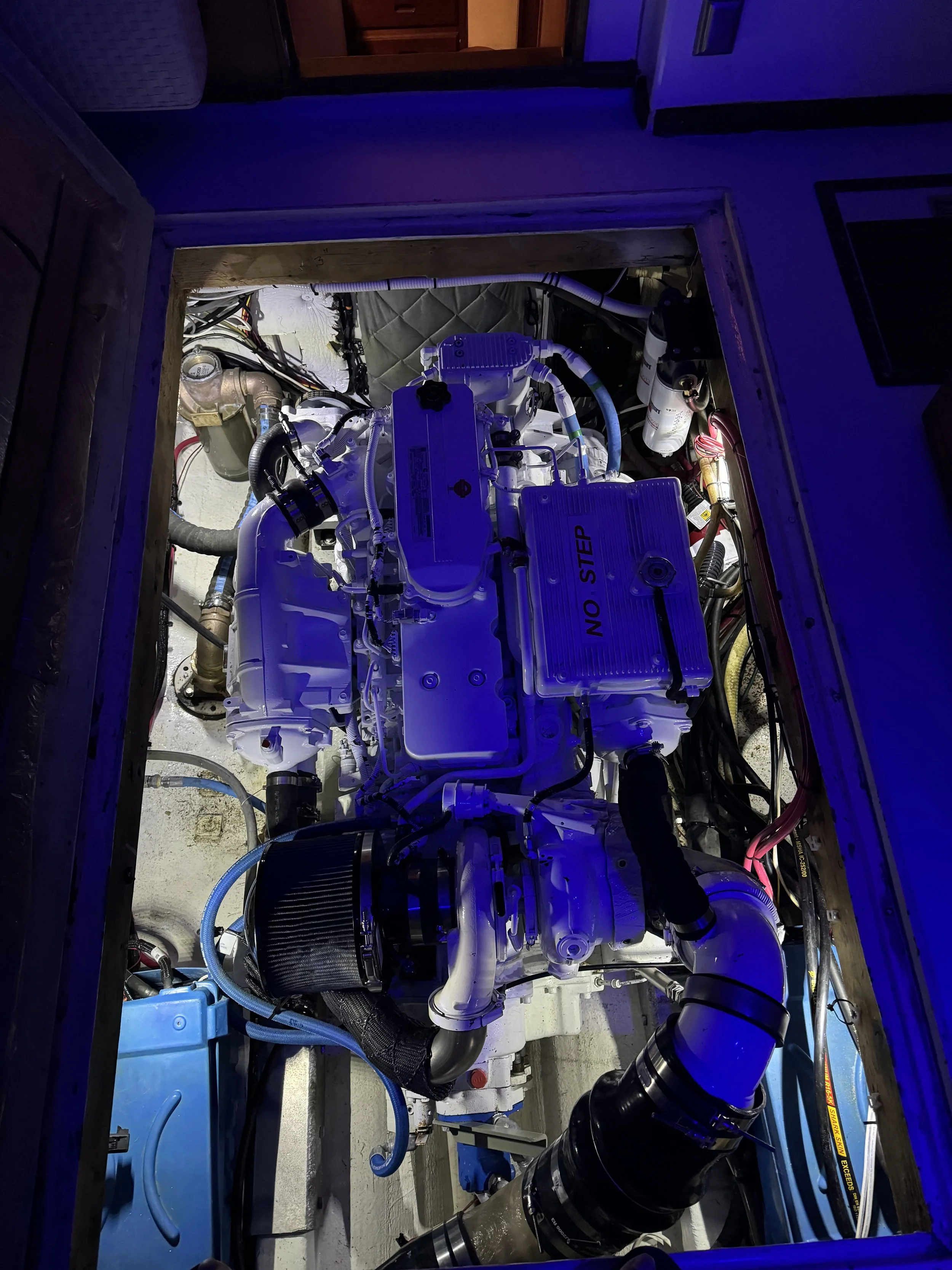 View of a boat engine compartment with a white marine engine labeled 'NO STEP,' surrounded by black and white hoses, wiring, filters, and marine equipment, illuminated by blue light.