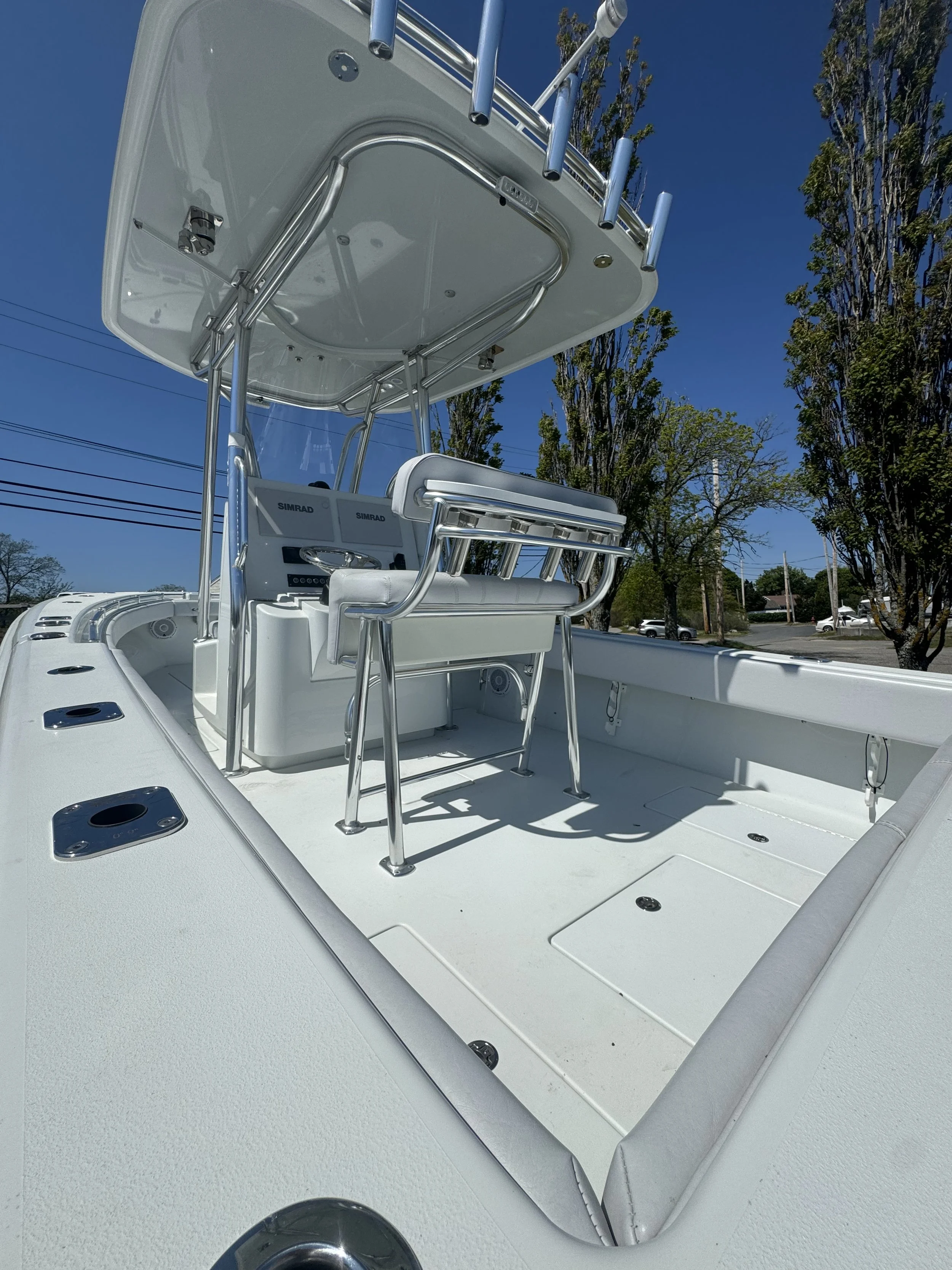 A white fishing boat with a T-top and multiple rod holders, stored on land with trees and power lines in the background.