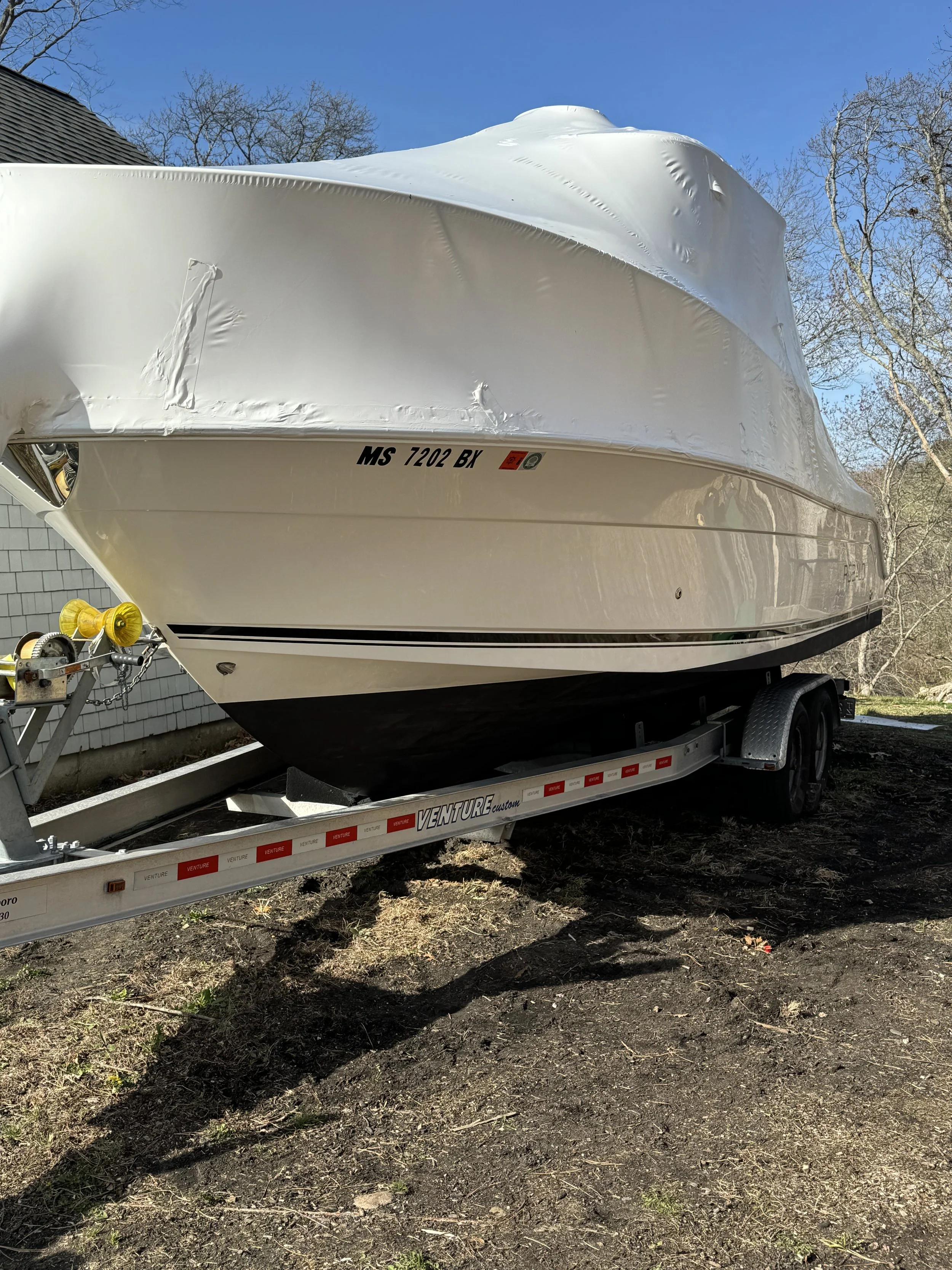 A white boat on a trailer parked outdoors on dirt, with a blue sky and leafless trees in the background.