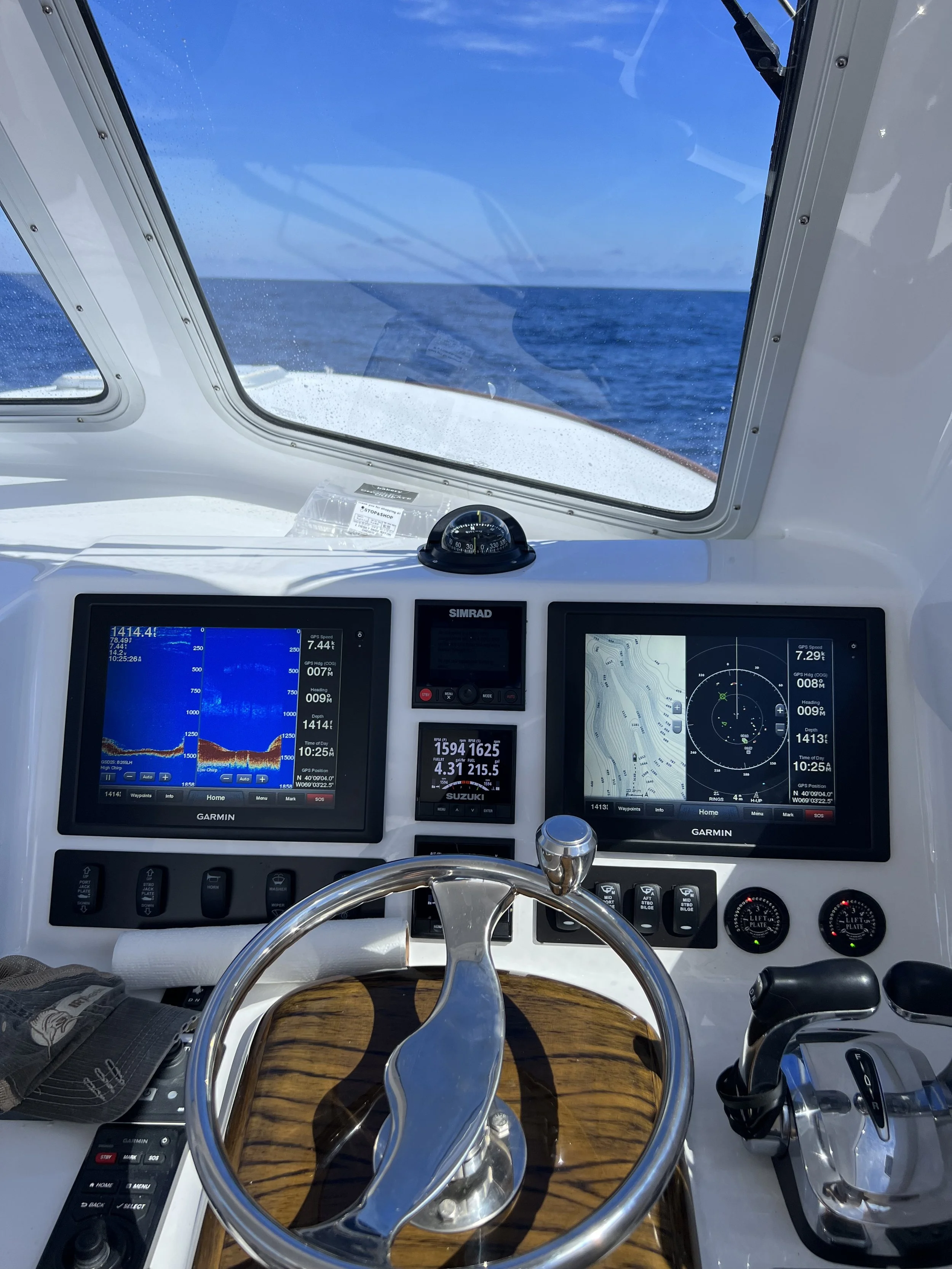 View from the helm of a boat showing navigational instruments, a compass, and a steering wheel, with the ocean and blue sky ahead.