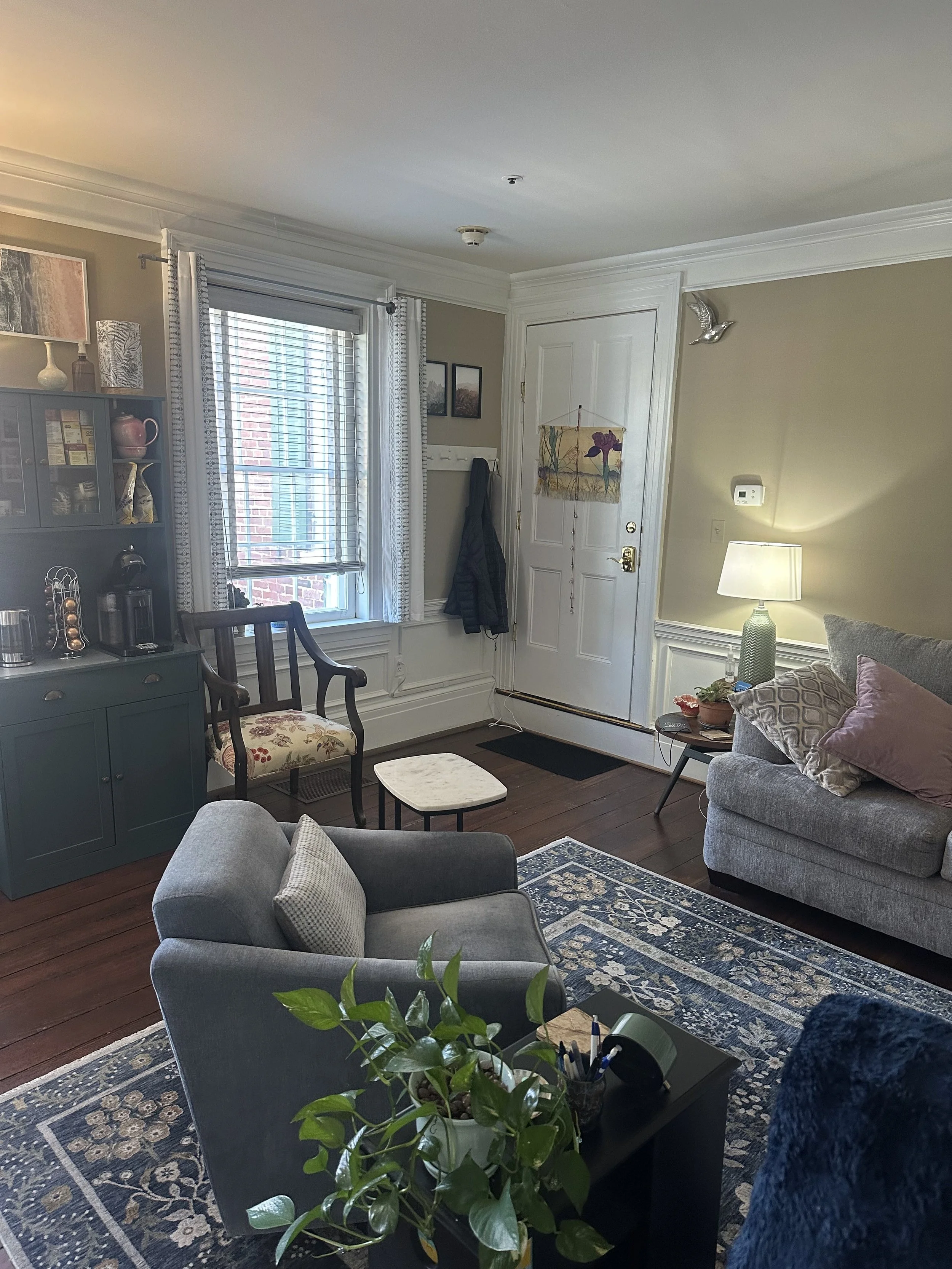 A picture of JJC Psychotherapy's Fairfax, VA office with a gray couch, floral armchair, wooden flooring, a window with white blinds and curtains,  wall art, a small table with a lamp, a coat hanging by the door, and a patterned rug.