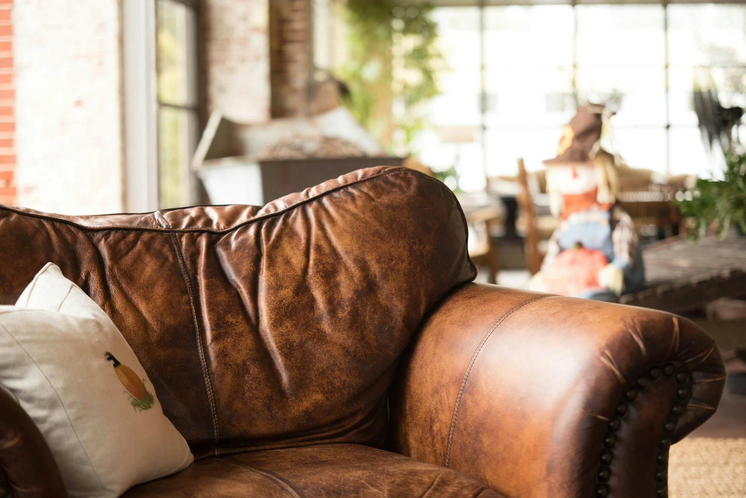 Close-up of a brown leather sofa with a white pillow on the left side, in a sunlit living room with large windows, trees, and a person sitting in the background.