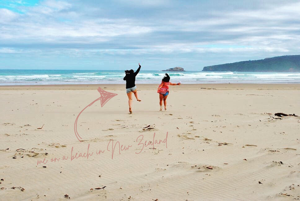Two women are running on a sandy beach towards the ocean with waves and a distant island or landmass in the background. One is wearing a black hoodie and shorts, and the other is in a red patterned top and shorts. There is a handwritten note and arrow pointing to the woman in the black hoodie, stating "me on a beach in New Zealand."