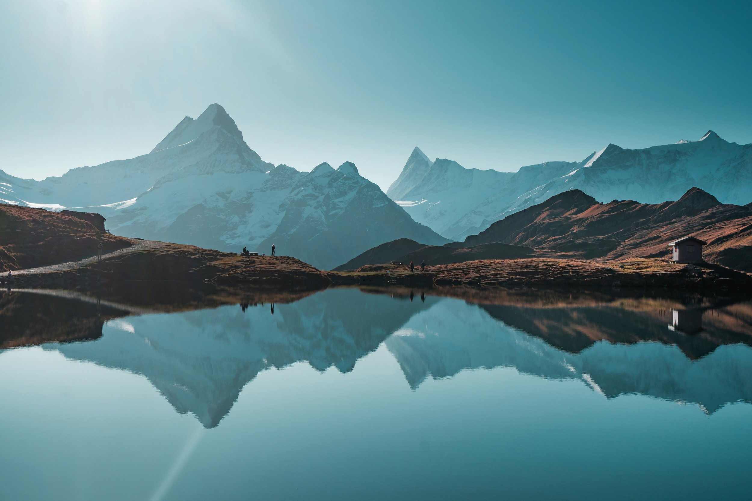 Snow-capped mountains reflecting in a calm lake with grassy hills and a small house. Silhouetted people walking on the hillside.