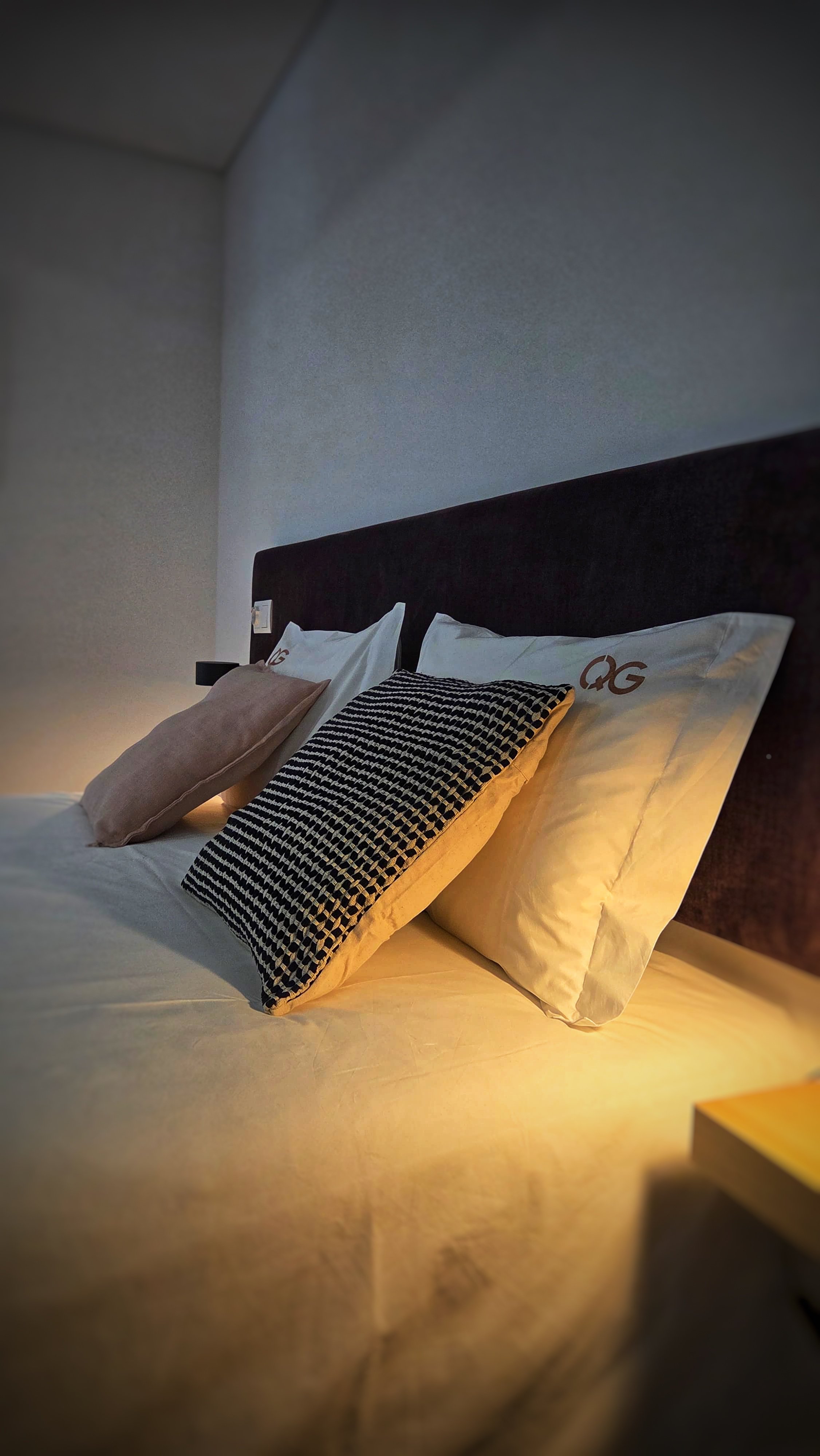 Lits d'hôtel avec plusieurs coussins, draps blancs, tête de lit en tissu noir et éclairage tamisé.