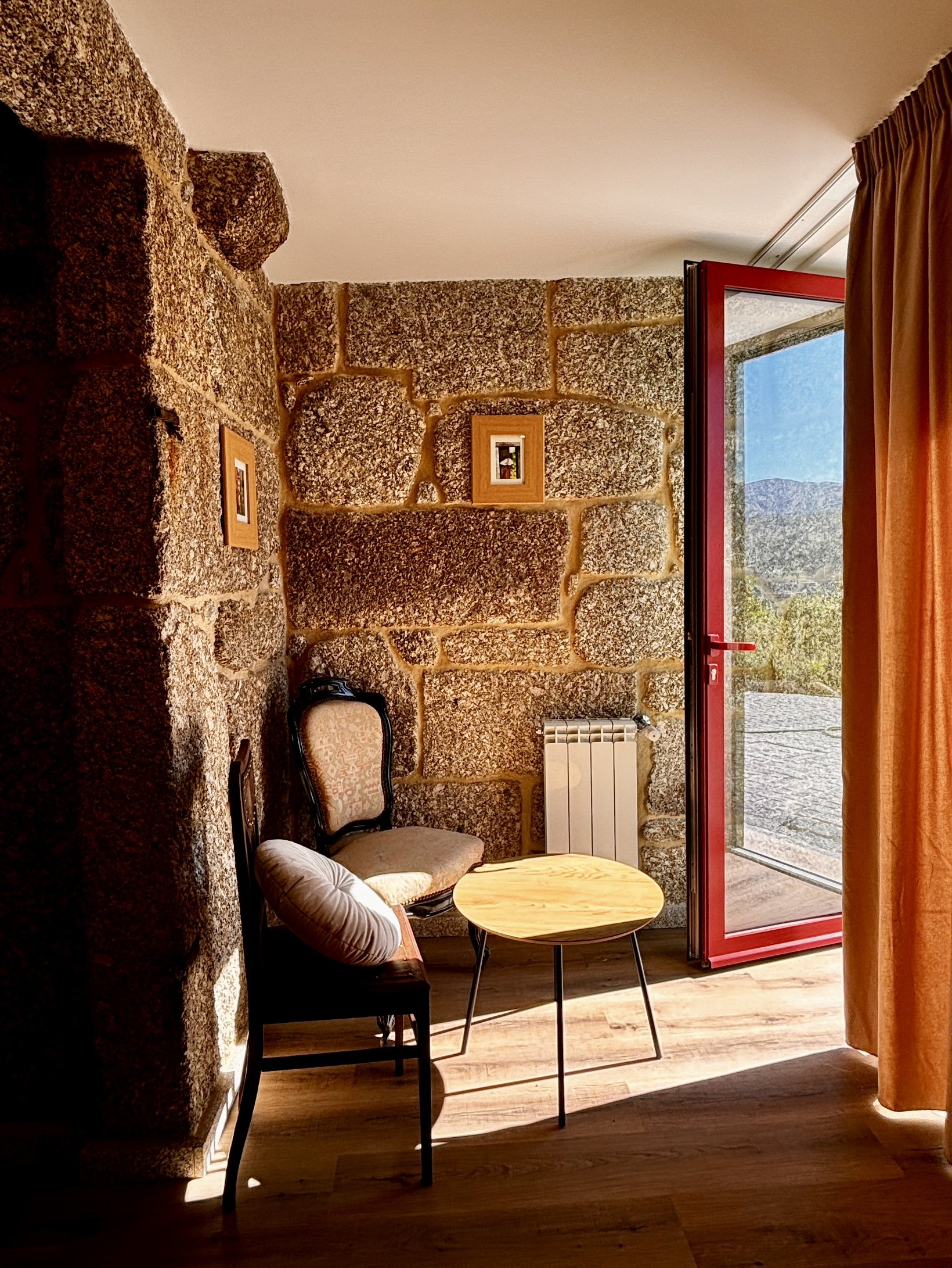 Une pièce with une fenêtre ouverte donnant sur un paysage montagneux, mobilier comprenant deux chaises, un coussin rond, et une petite table en bois, mur en pierre