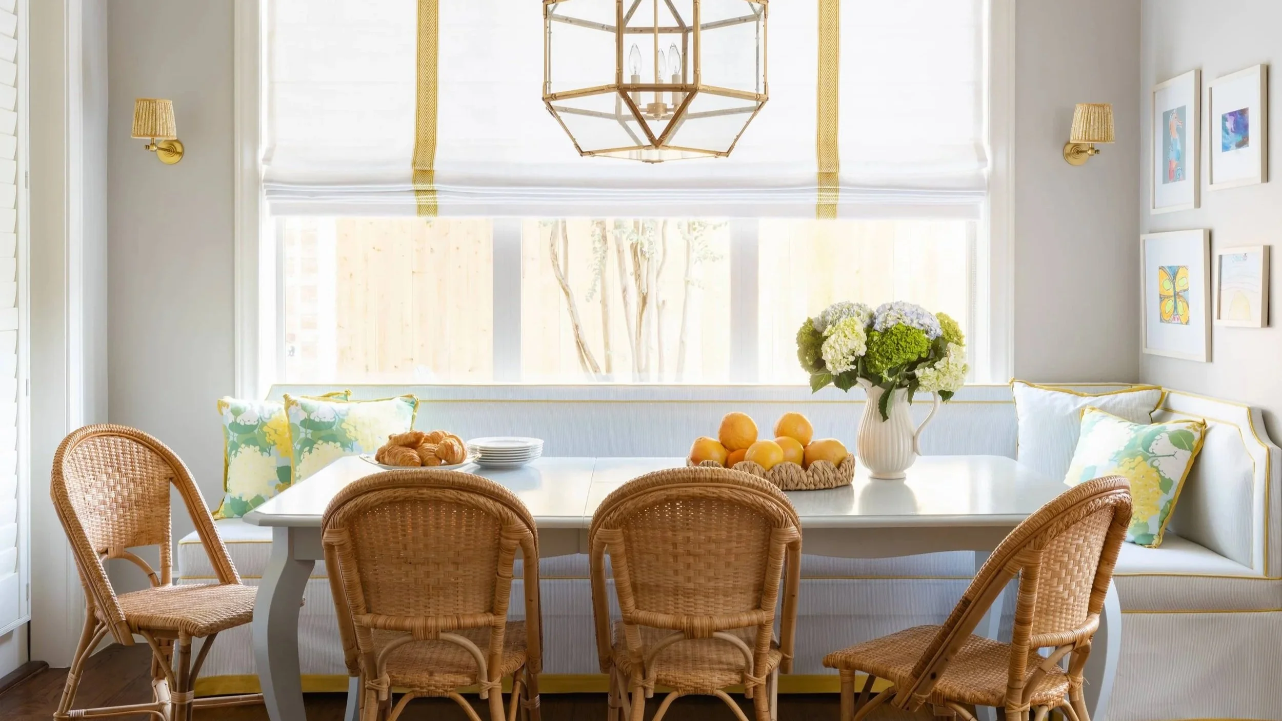 Bright dining area with a white table surrounded by woven rattan chairs, adorned with floral pillows. The table holds a vase with white and green hydrangeas, a basket of yellow fruits, croissants, and a plate. Large windows with white shades allow natural light, and a geometric chandelier hangs above. Framed colorful artwork decorates the white wall, with gold wall sconces flanking the window.