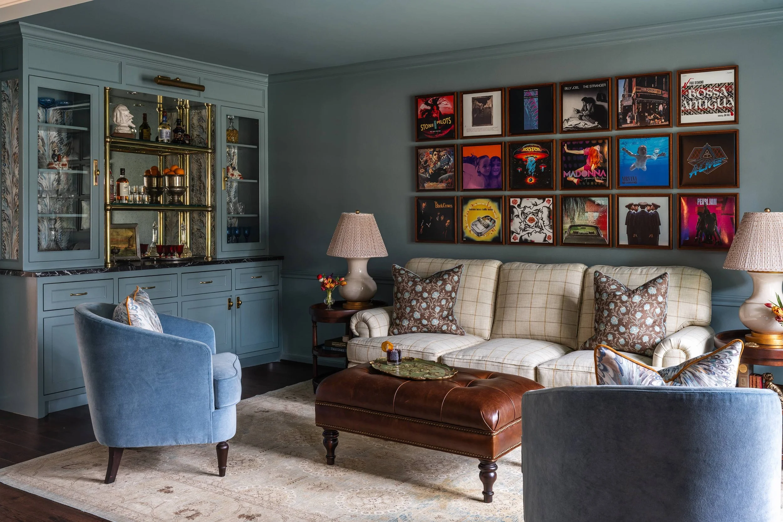 Living room with light blue walls, a beige checkered sofa, blue armchair, and a wooden coffee table. Framed album covers adorn the wall above the sofa. There is a blue cabinet with glass doors, gold accents, and a bar with bottles and glasses. Two table lamps with beige shades sit on side tables, and a beige area rug covers part of the dark wood floor.