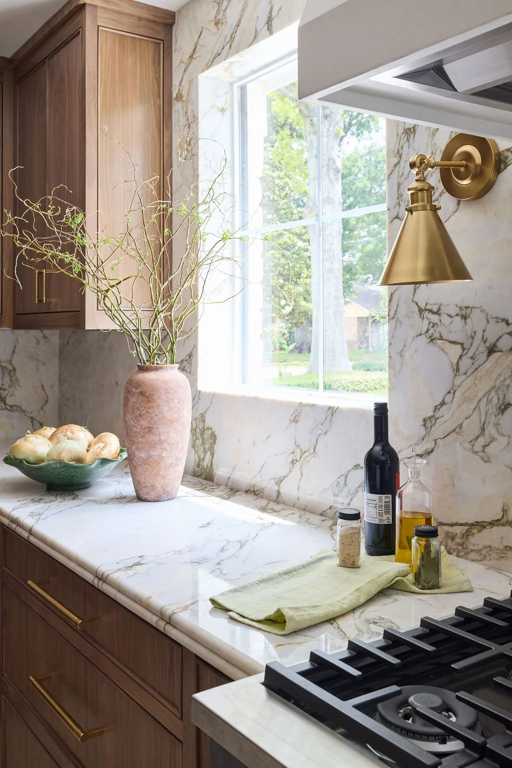 A kitchen counter with a beige marble surface, a vase with curly twigs near a window, a bowl of onions, wine bottles, oil, spices, a green towel, and a gas stove, with a large window showing trees outside. Beautiful kitchen interior design in Houston