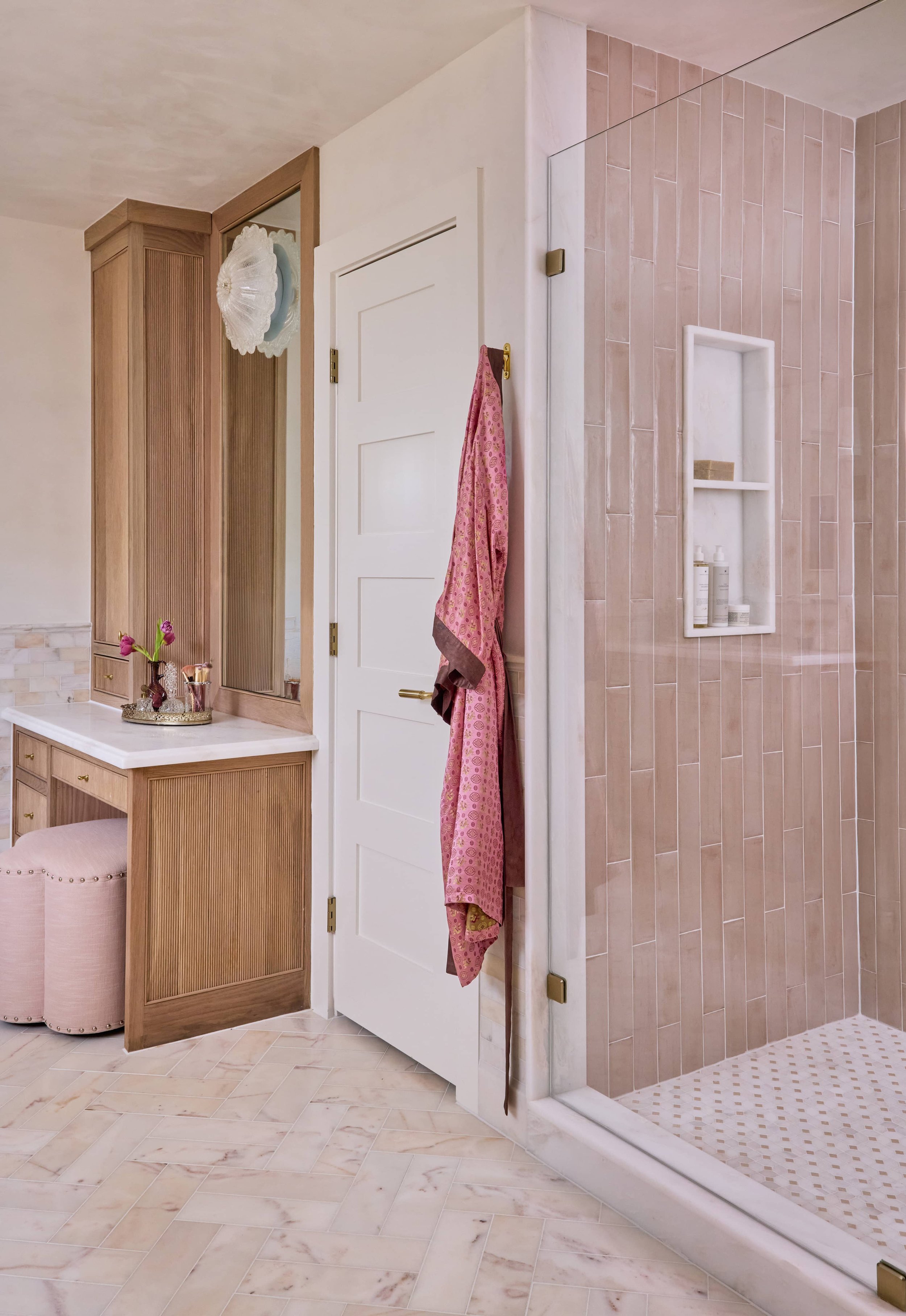A bathroom with a tiled shower area, a pink towel hanging, a wooden cabinet with a glass door, a beige cushioned stool, and a decorative tray with flowers on a marble countertop. Beautiful bathroom interior design in Houston, Texas.