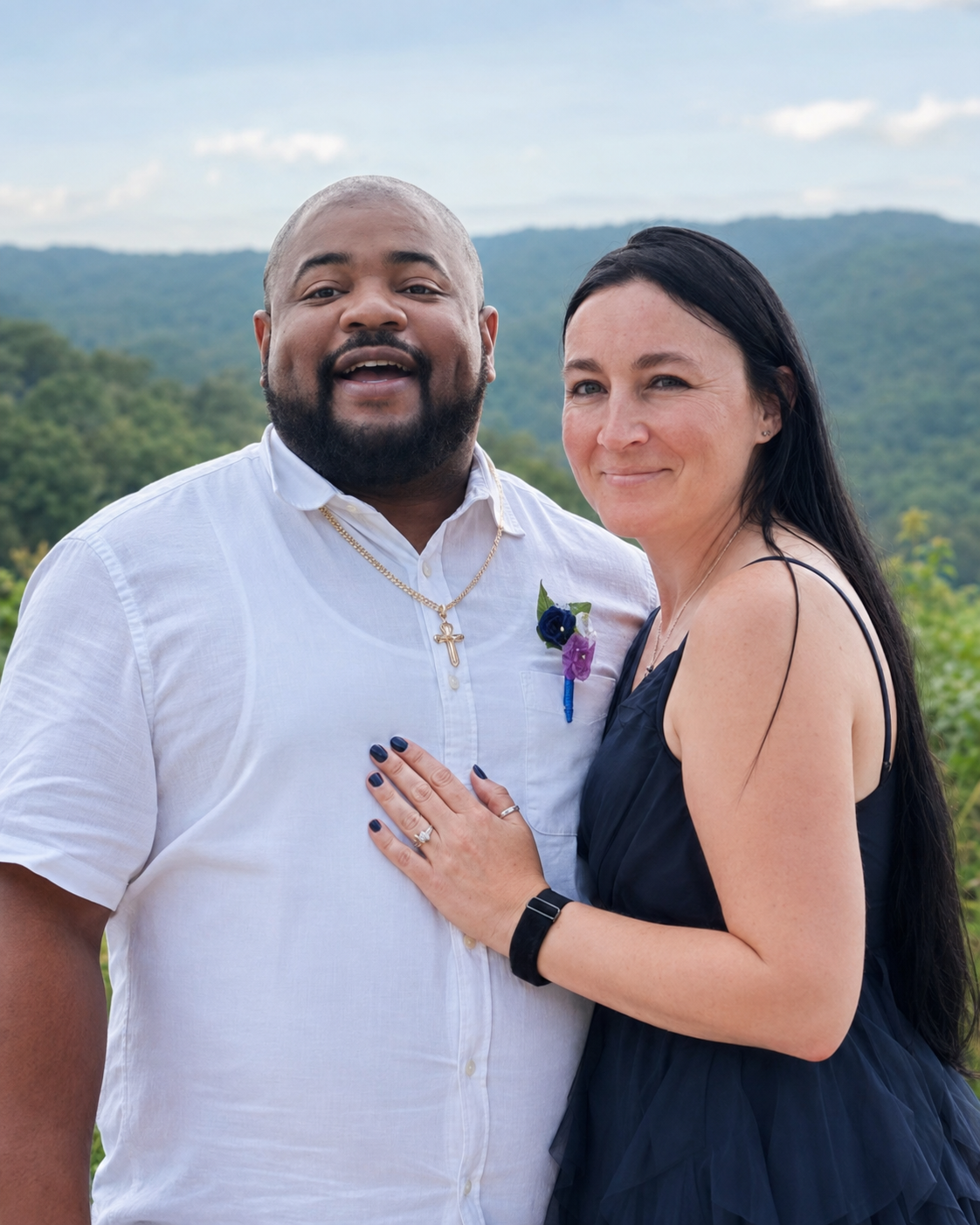 Happy couple standing outdoors with mountains and cloudy sky in the background, woman wearing a black dress and wedding ring, man wearing a white shirt, gold chain, and boutonniere.