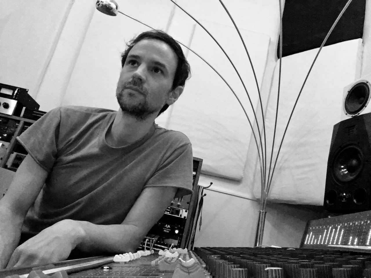 A man with dark hair, a beard, and wearing a t-shirt sitting in a music studio or control room with audio equipment and speakers in the background.