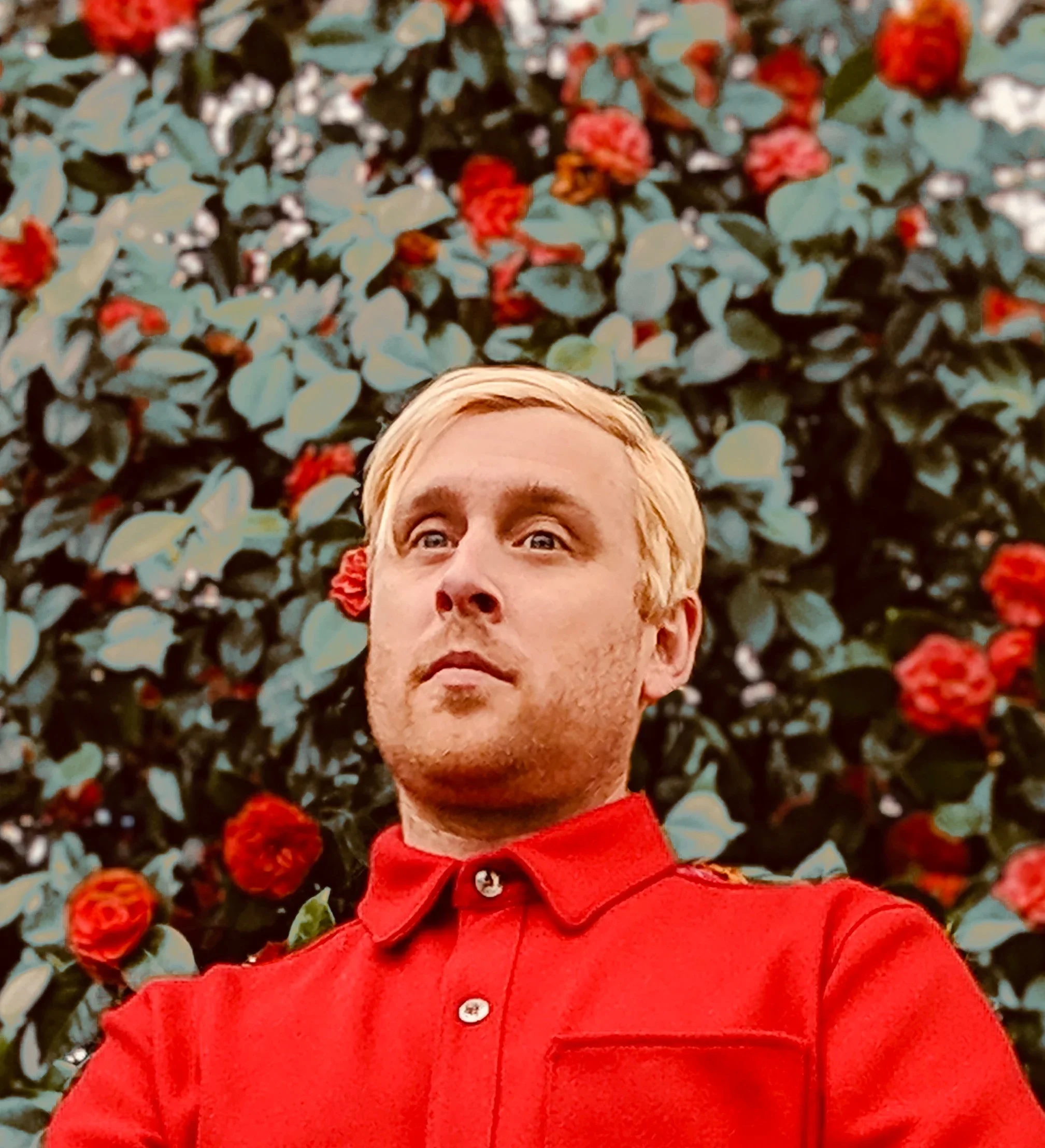 A smiling man with glasses and short blond hair, wearing a green T-shirt with a cartoon character, is working with audio or electronic equipment in a recording studio.