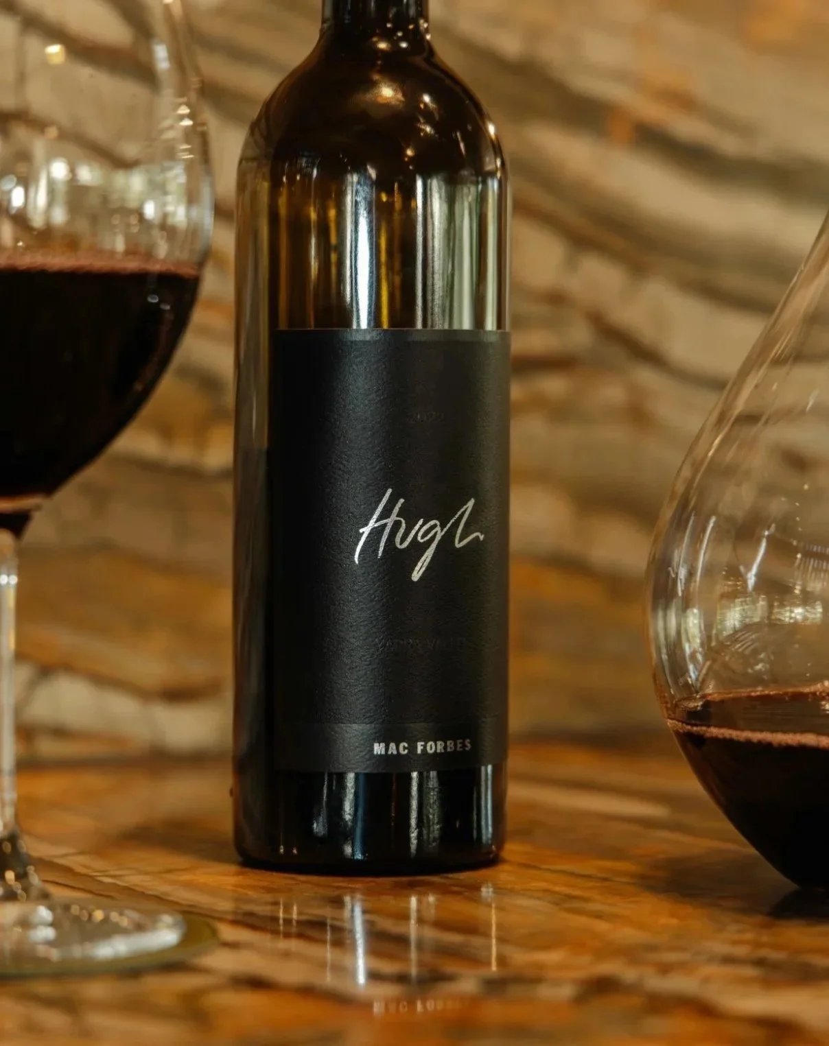 A black wine bottle labeled 'Hugh' placed on a wooden table with two wine glasses filled with red wine, one on each side of the bottle.