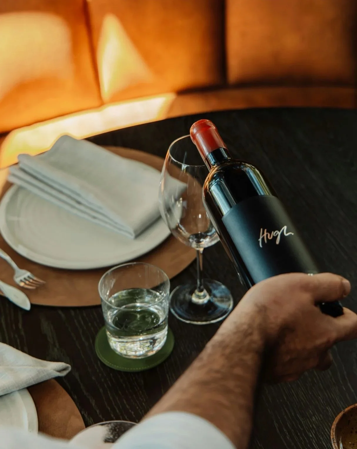 A person pouring red wine into a glass on a dining table set with a plate, silverware, and folded napkins.