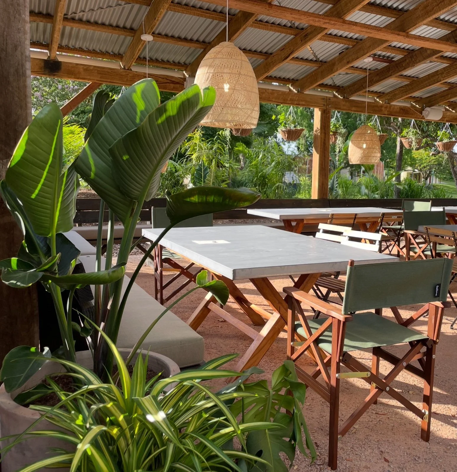 Outdoor dining area with wooden tables and green chairs under a wooden roof, surrounded by lush green plants and hanging wicker lamps.