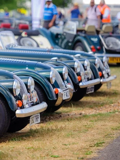 Classic green vintage cars lined up outdoors at a car show with people walking in the background.
