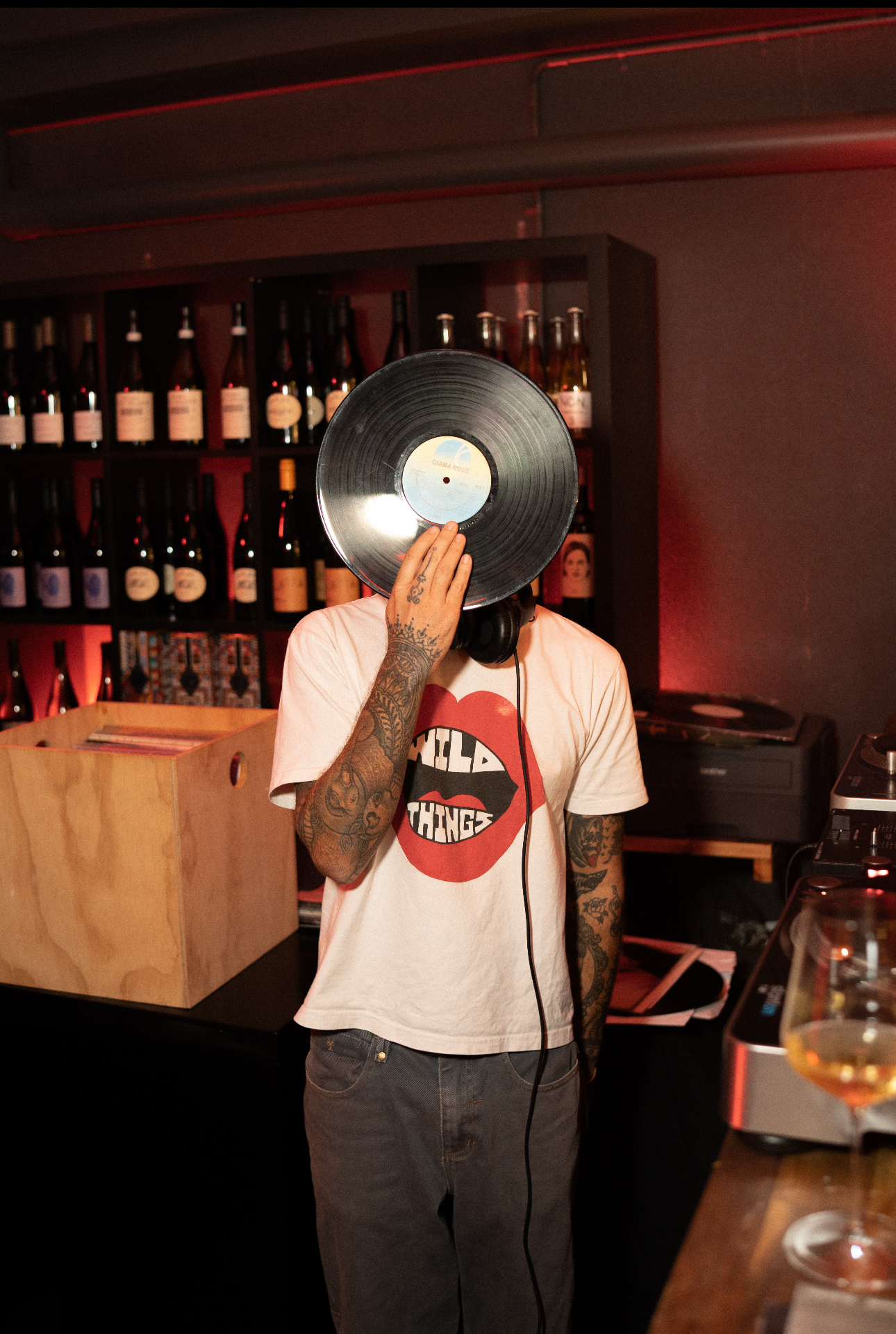 A person with a beige apron is preparing food at a rustic kitchen counter, holding a small bowl in one hand and drizzling a dark sauce over roasted vegetables on a plate with a spoon in the other hand.