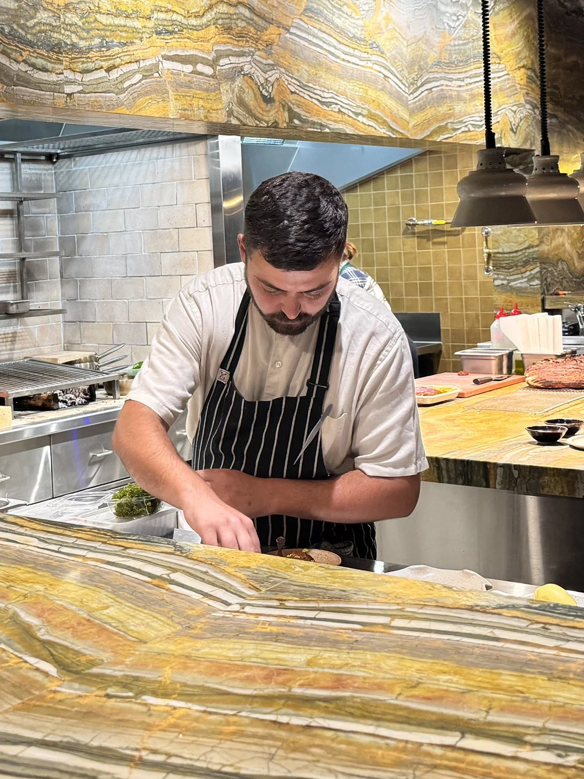 A chef in a striped apron preparing food at a marble countertop in a restaurant kitchen.