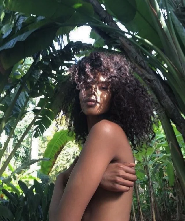 A woman with curly hair standing among lush green tropical plants, covering her chest with one arm.