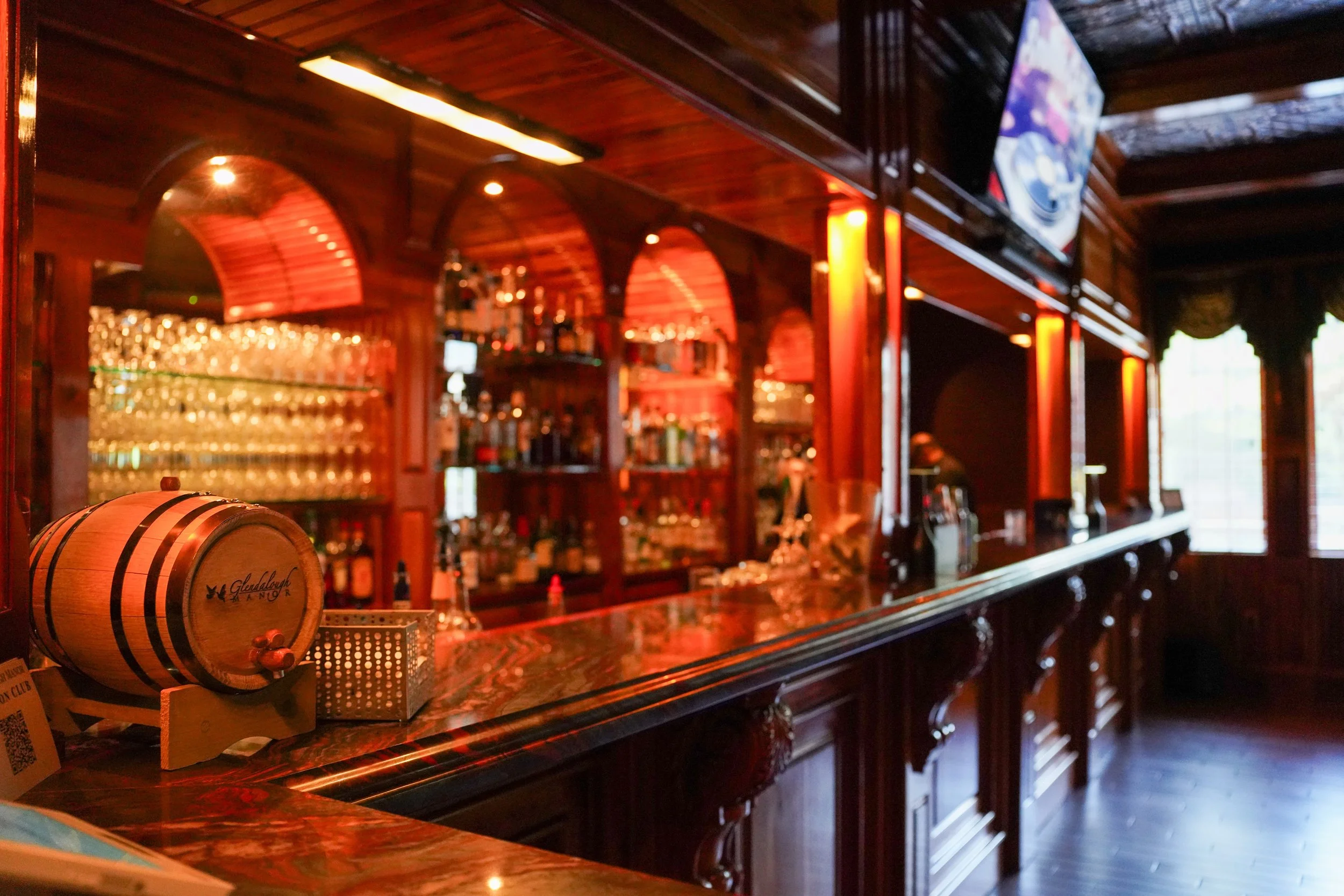 Interior of a bar with wooden counters and shelves, a small barrel on the counter, and televisions mounted on the wall.