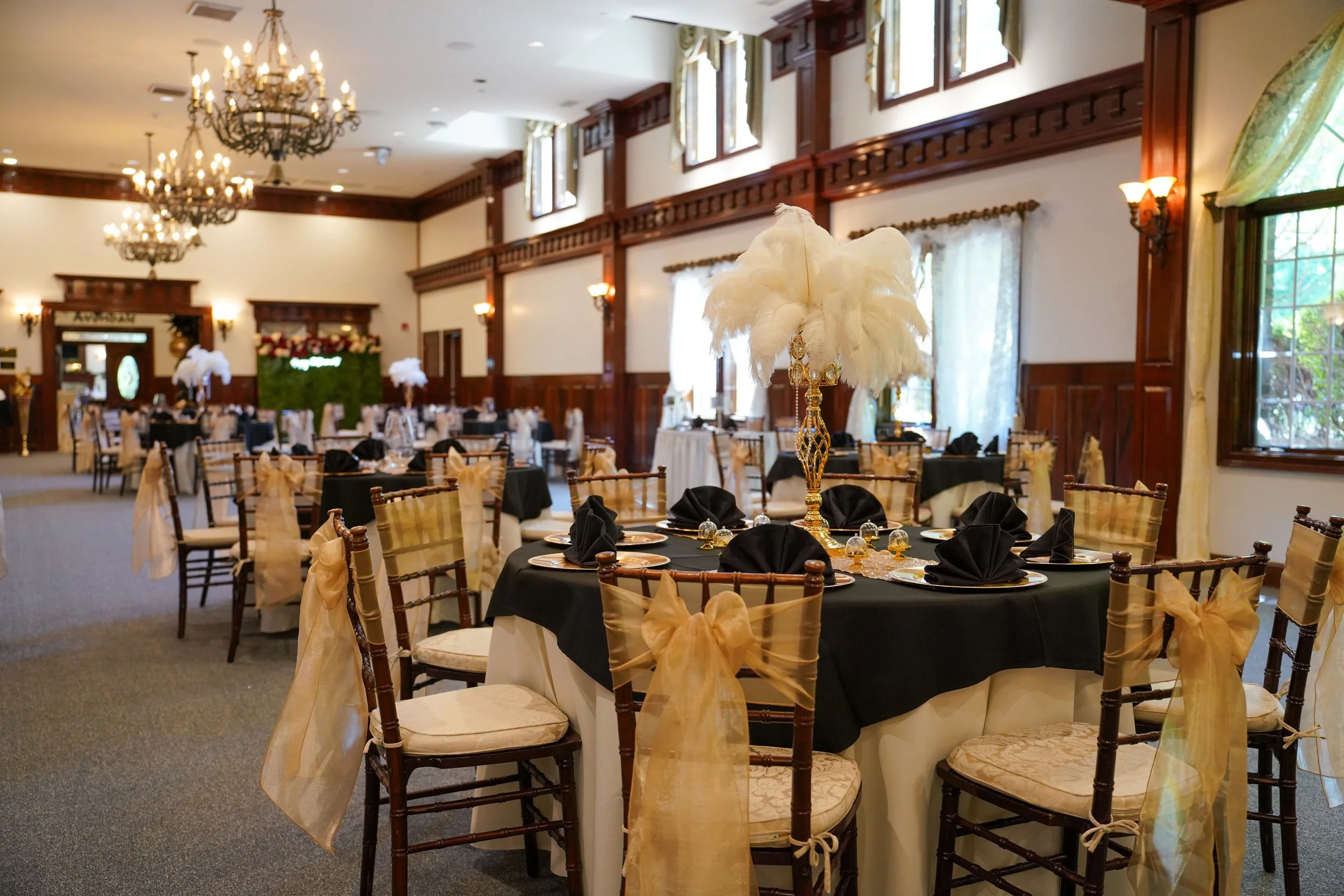 Elegant banquet hall decorated for event with round tables covered in black cloth, gold bows on chairs, and tall centerpieces with white feathery decorations, chandeliers hanging from ceiling, large windows with curtains, and wood paneling on walls.