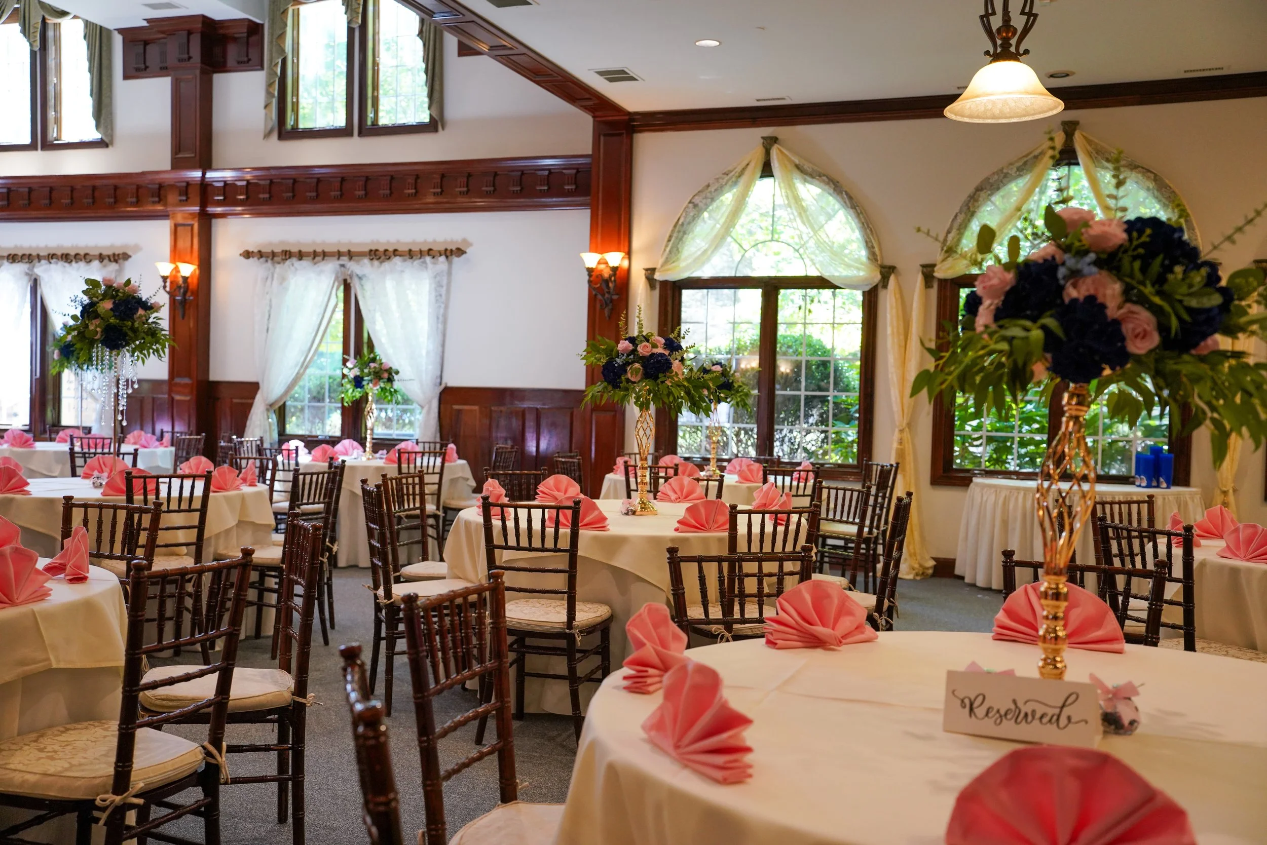 A banquet hall decorated for an event with round tables covered in white tablecloths, pink napkins, floral centerpieces, and reserved signs. The hall has large windows with sheer curtains, wood paneling, and a mix of wall sconces and a hanging light 