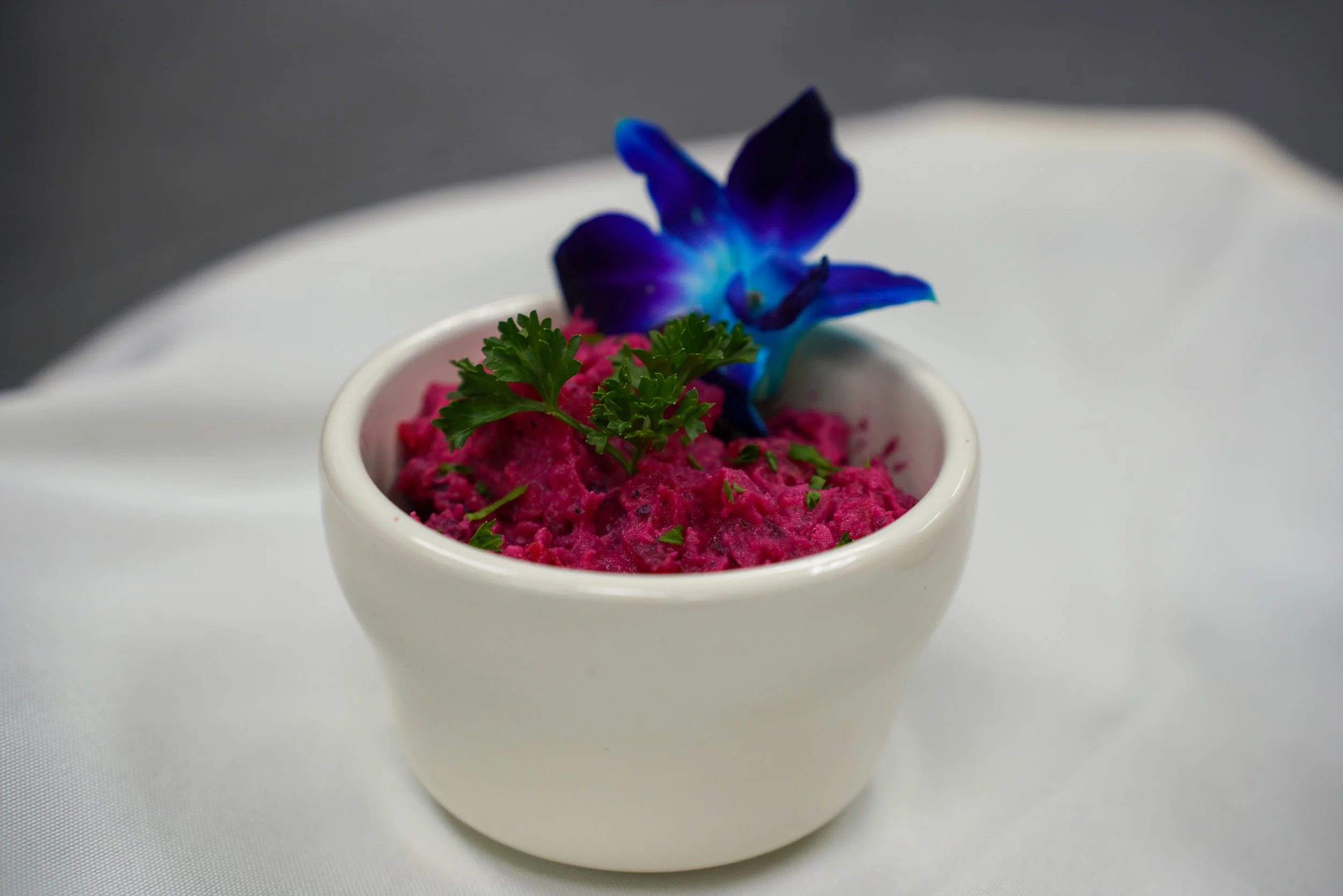 A small white bowl filled with bright pinkly colored mashed sweet potatoes, garnished with a sprig of parsley and a decorative blue flower.