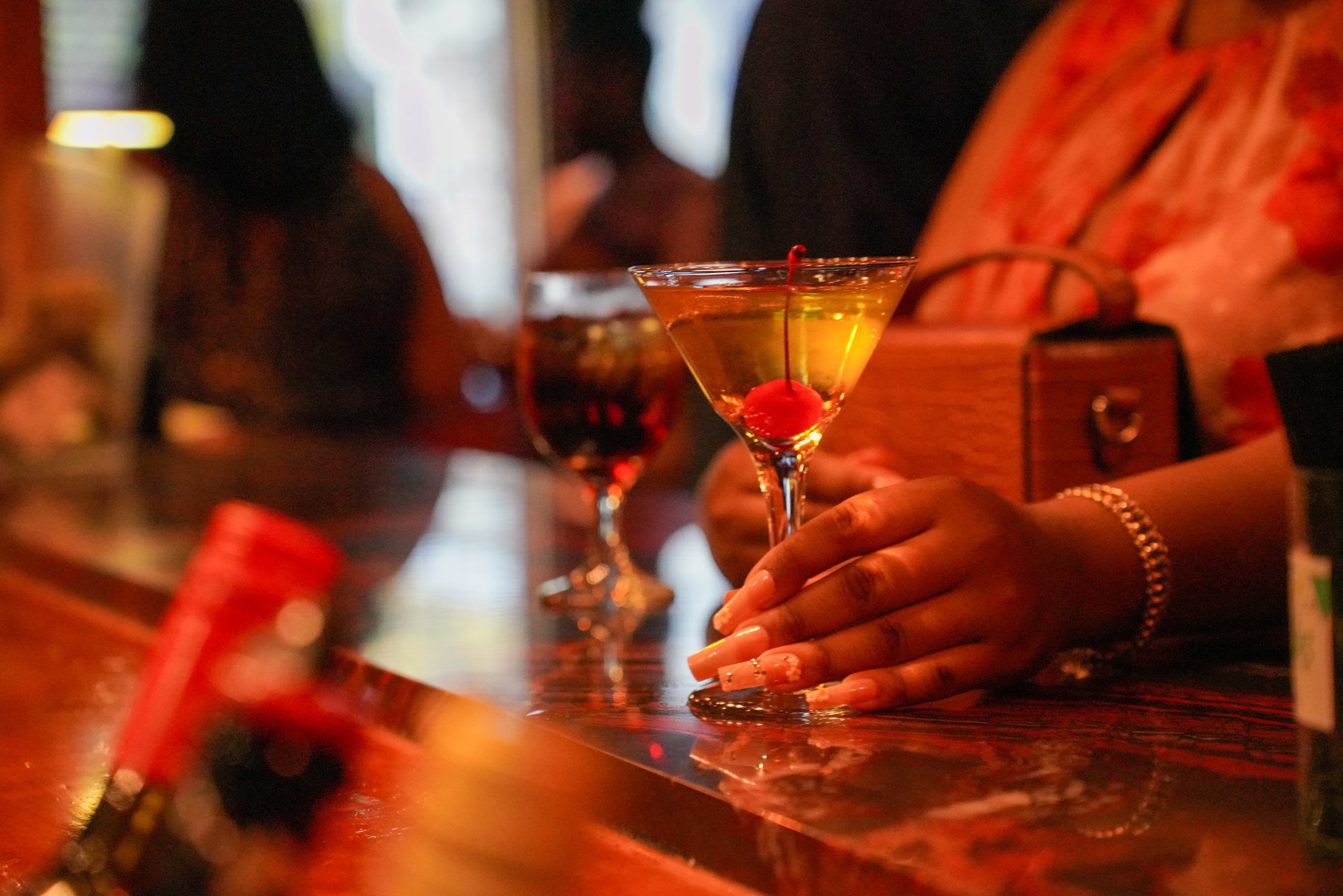 A hand with manicured nails holding a cocktail glass with a cherry garnish in a dimly lit bar or lounge.