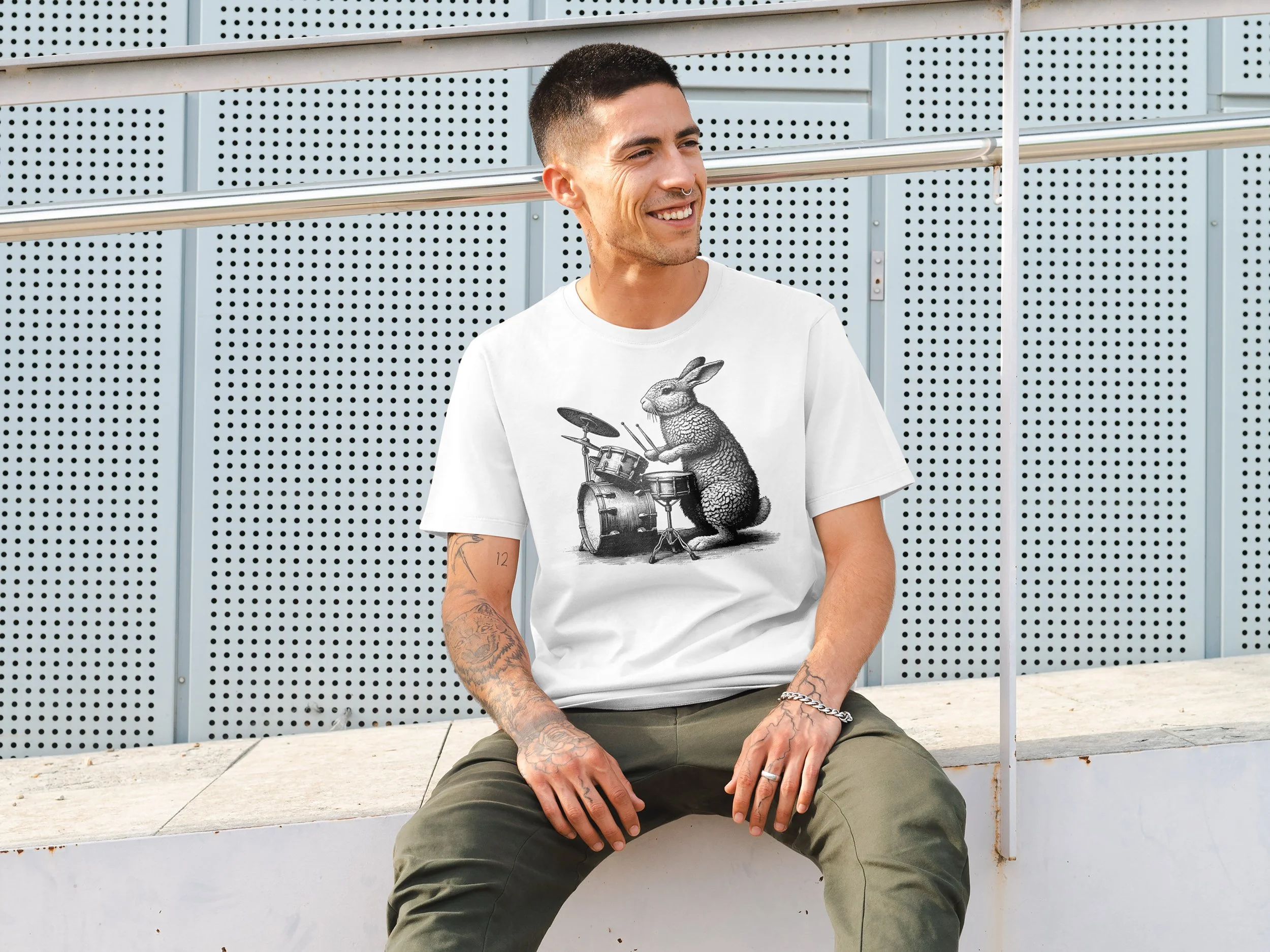 A young man sitting outdoors on a ledge, smiling and wearing a white t-shirt with a graphic of a rabbit playing drums, khaki pants, tattoos on his arms, and a bracelet.
