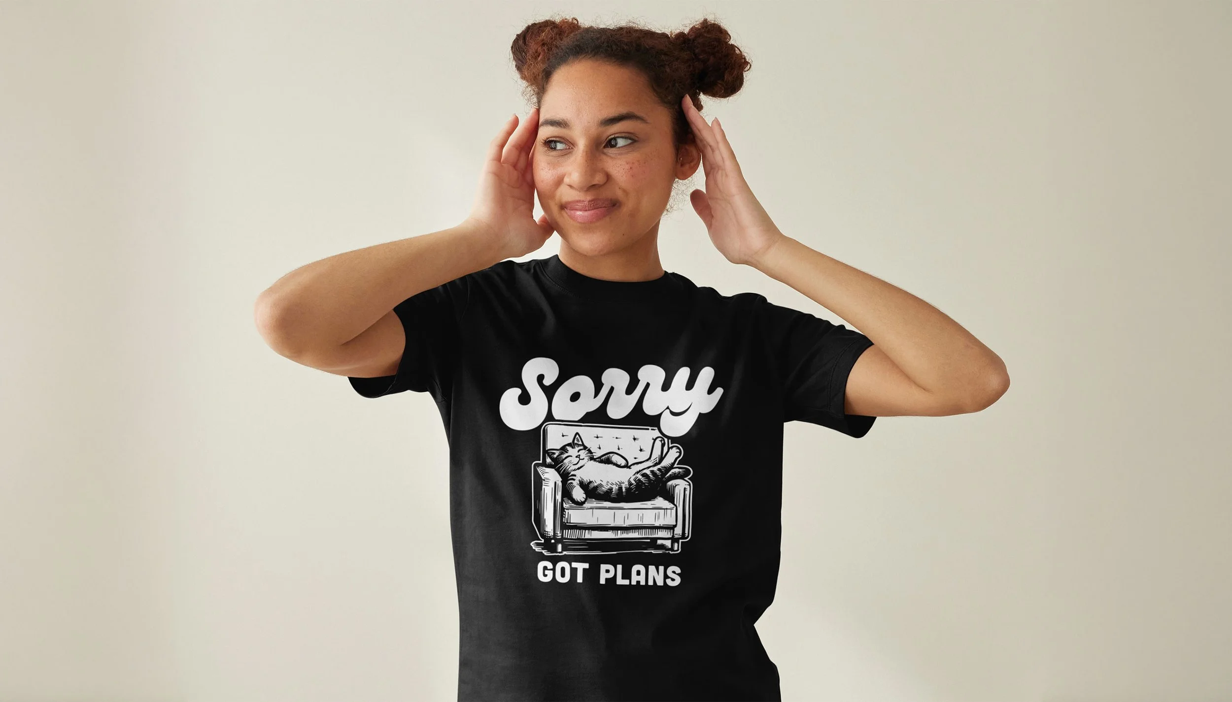 Young woman with curly hair holding her head, wearing a black t-shirt with a cat lounging on a couch and the text "Sorry Got Plans".